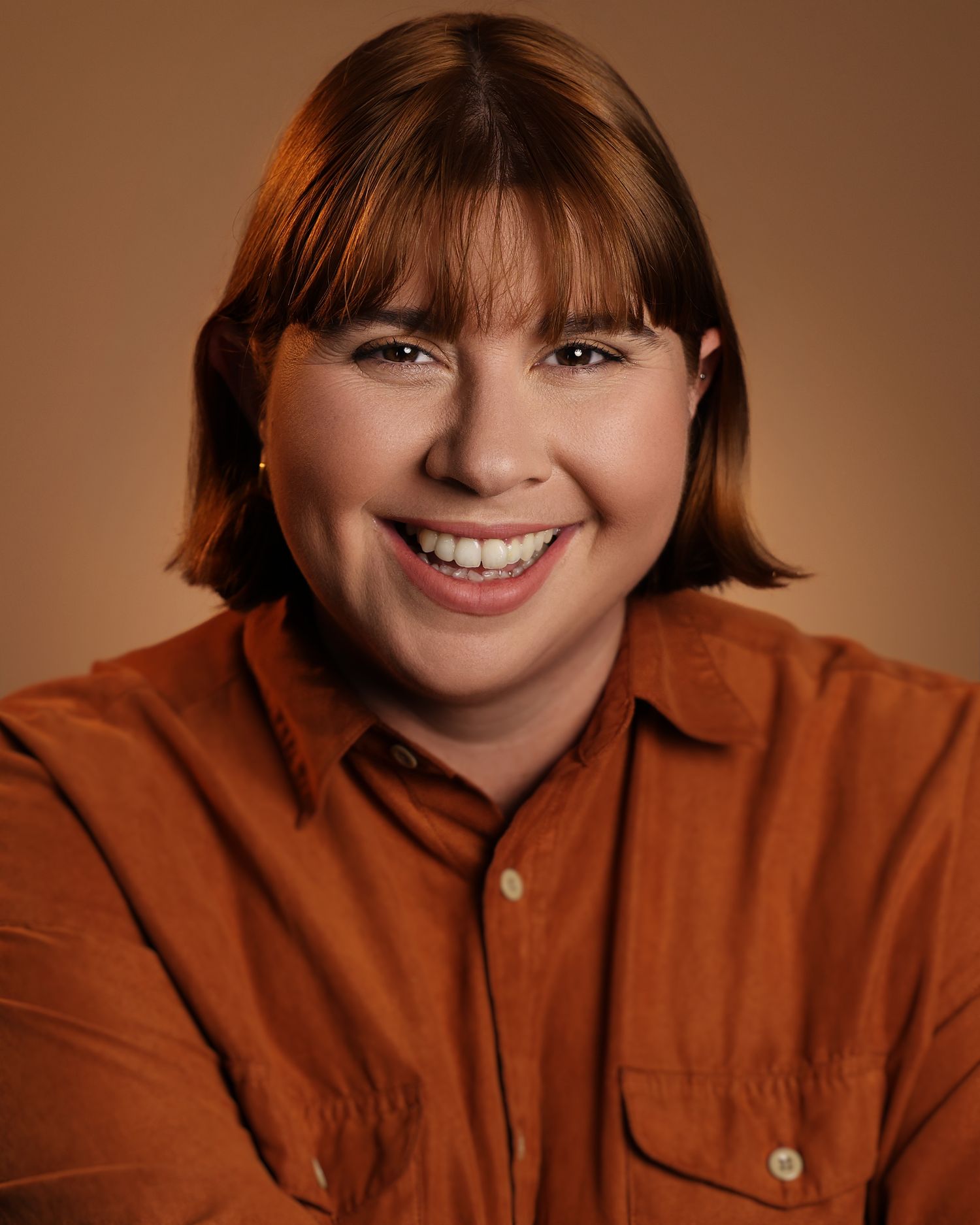 Smiling person with red bob haircut wearing an orange button-up shirt against a warm brown background.