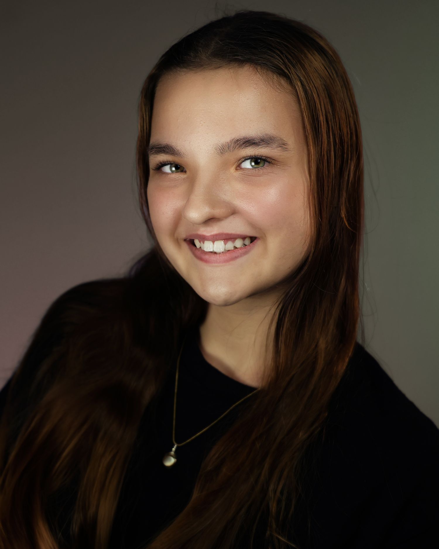 Smiling young woman with long brown hair wearing a black top and gold necklace against a gray background.