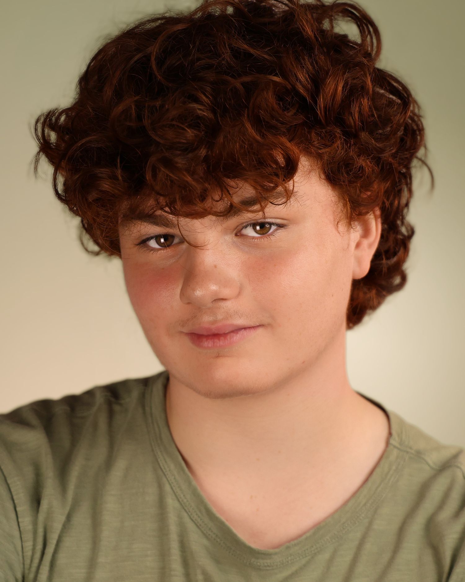 Young teen with curly red hair wearing a sage green t-shirt poses for a professional headshot photo.