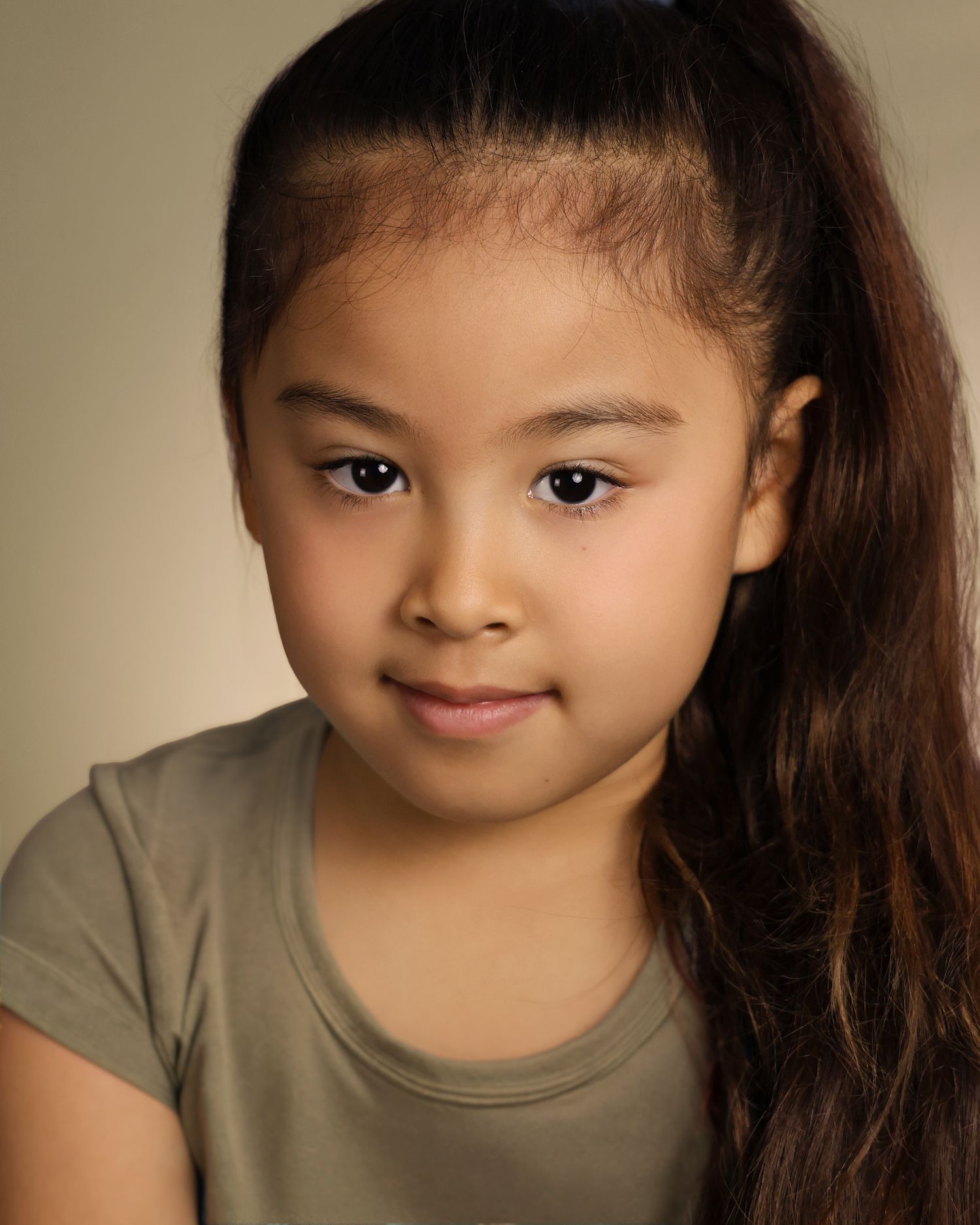 Young girl with long dark hair in a ponytail, wearing an olive green top, posing for a professional headshot.