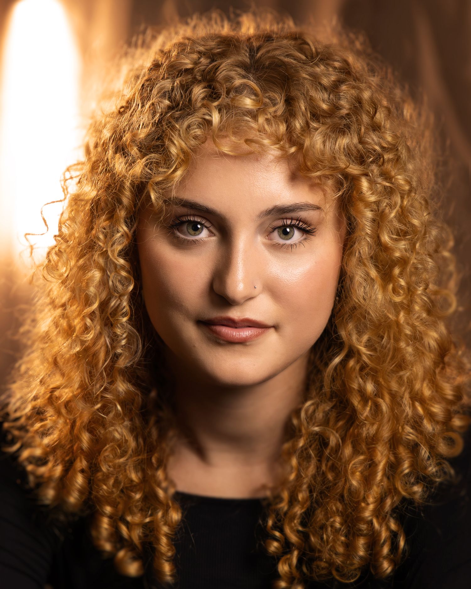 Young woman with voluminous golden curly hair poses for a professional headshot against a warm glowing background.