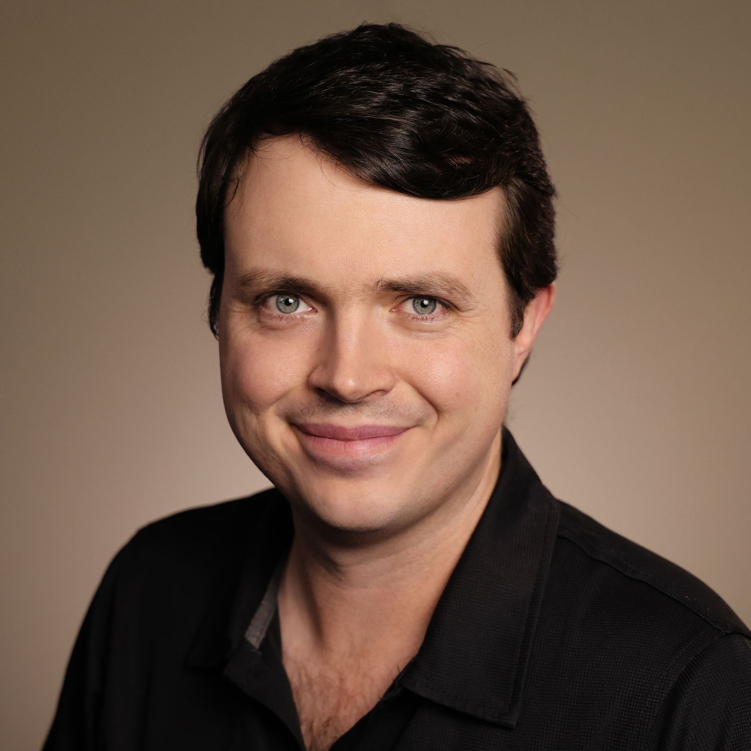 Professional headshot of a smiling young man with dark hair and blue eyes wearing a black shirt on a neutral background.