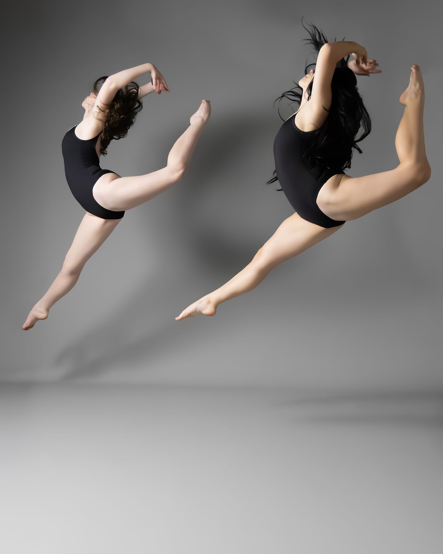Two female dancers leap gracefully in black leotards against a gray studio backdrop.