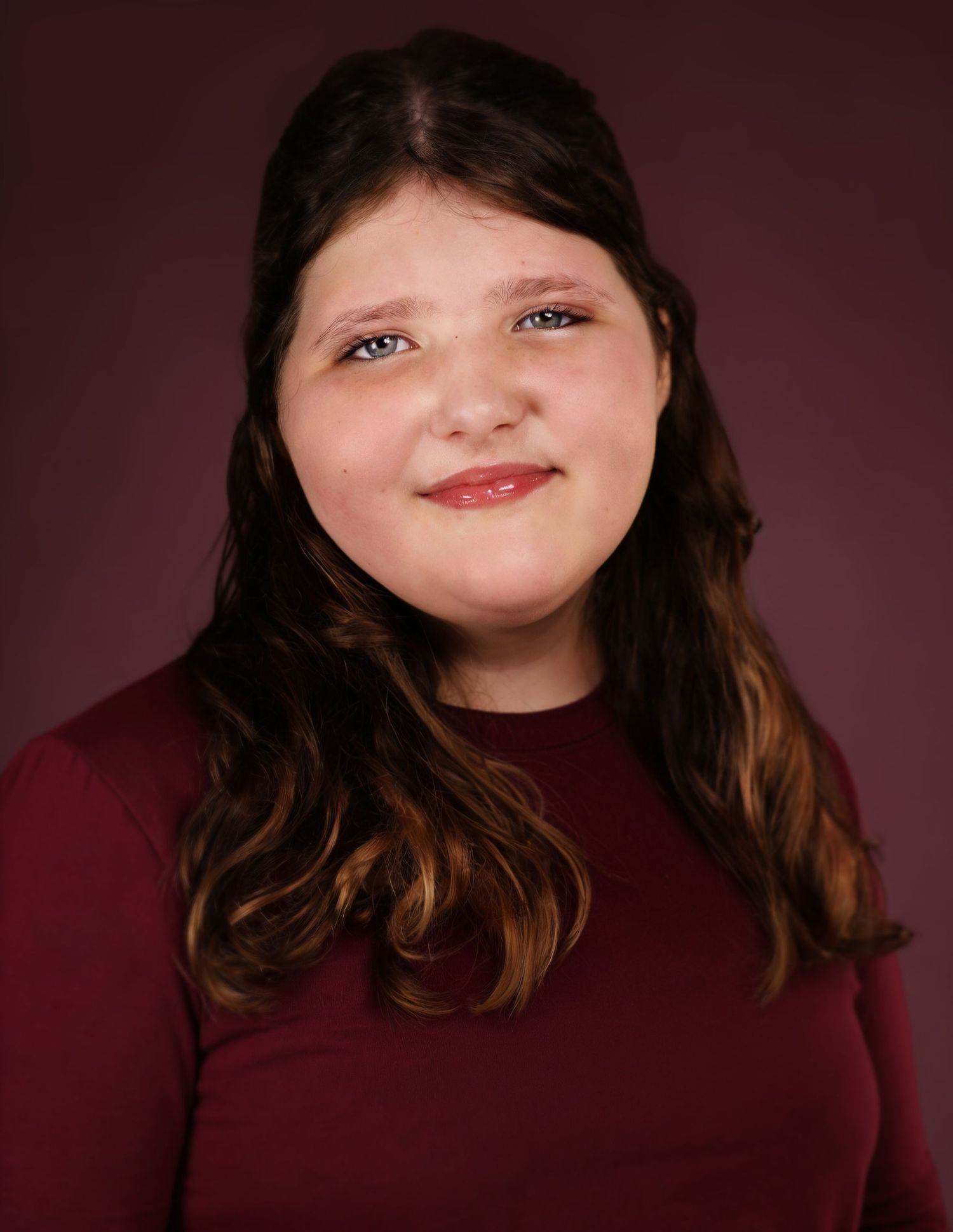 Young woman with long brown hair smiling, wearing a dark red top against a maroon studio background.