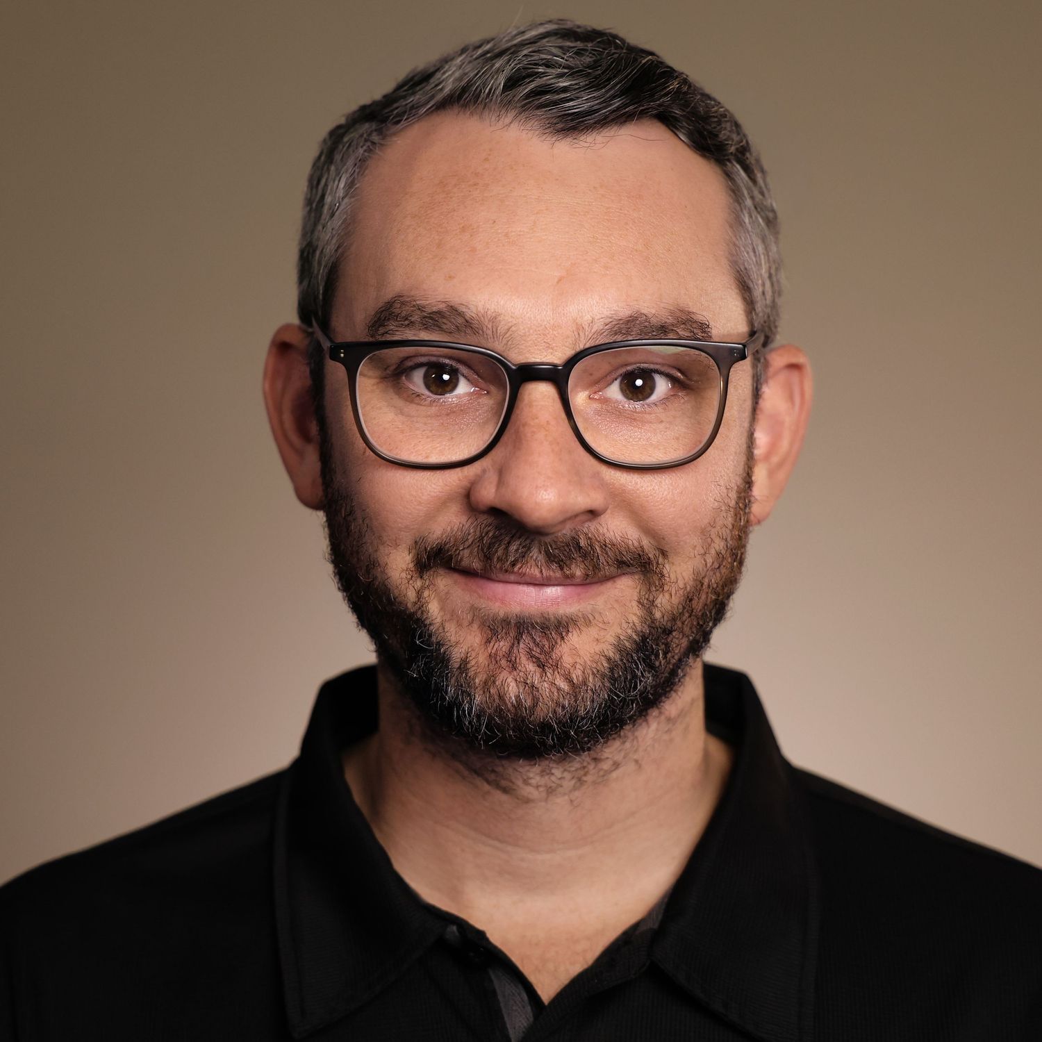 Professional headshot of a man with short gray hair, beard, and glasses wearing a black polo shirt.
