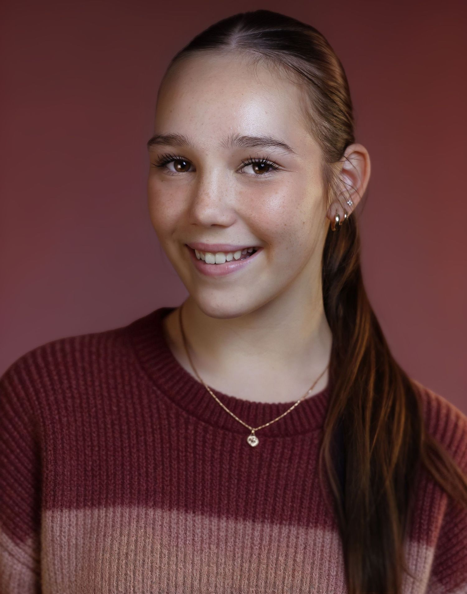 Smiling young girl with long brown hair, wearing a striped pink sweater and delicate gold necklace against a mauve background.