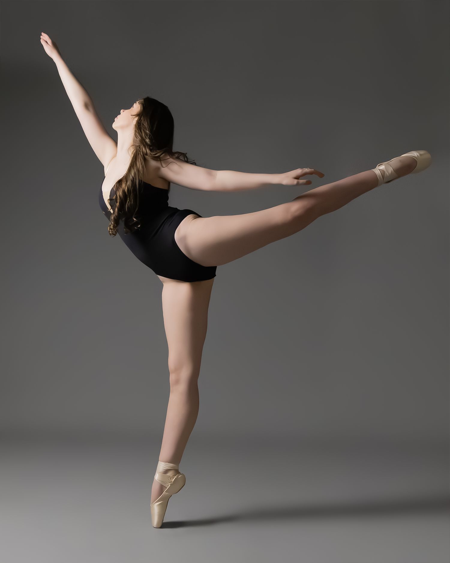A ballet dancer in a black leotard balances en pointe, extending one leg high while raising her arm gracefully.