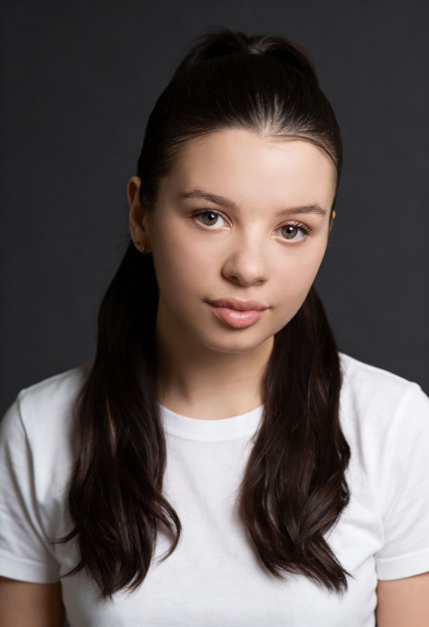 Young woman with dark hair in a half-up style wearing a white t-shirt against a gray background.