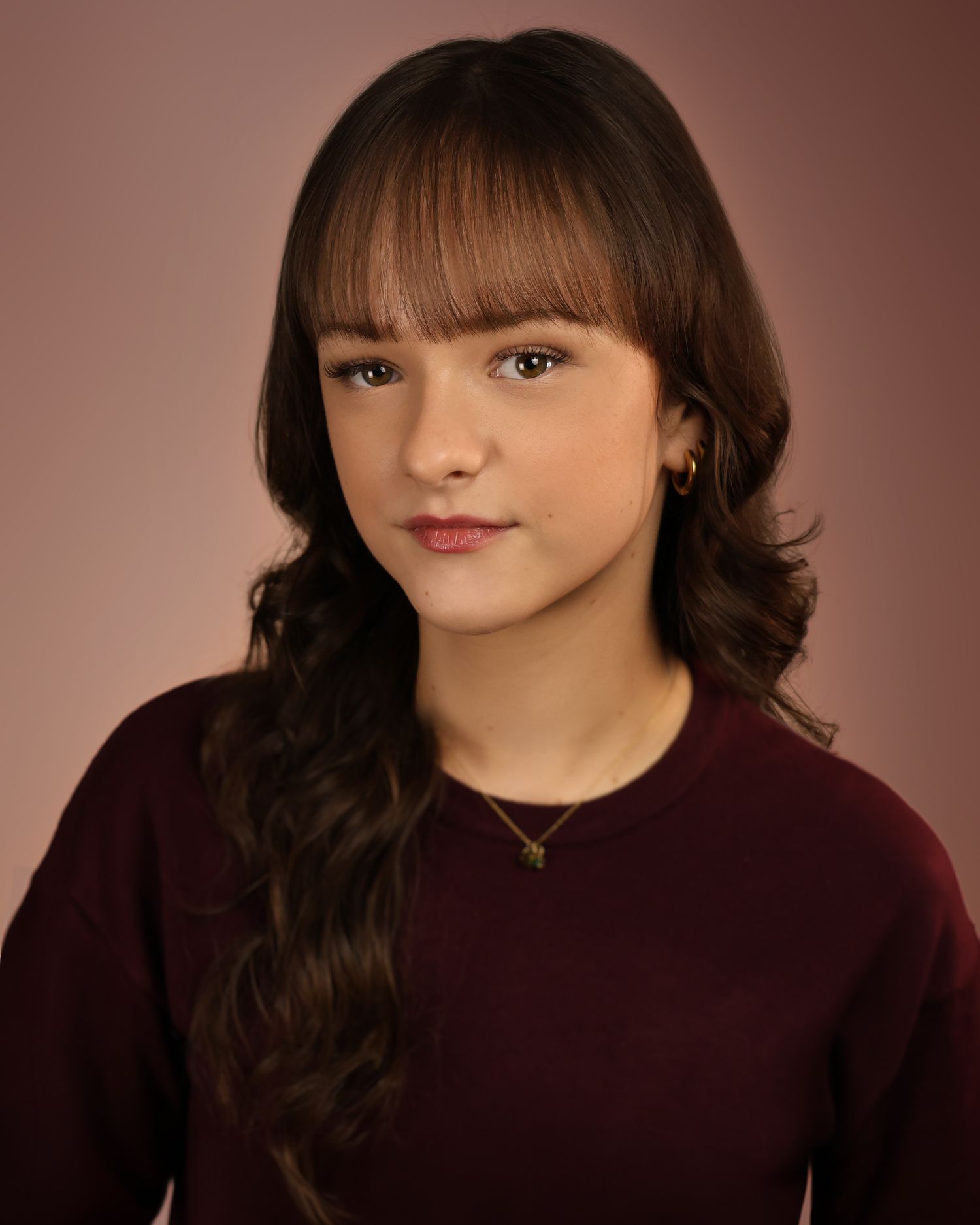 Young girl with dark wavy hair and bangs wearing a burgundy top, posing for a professional headshot photo.
