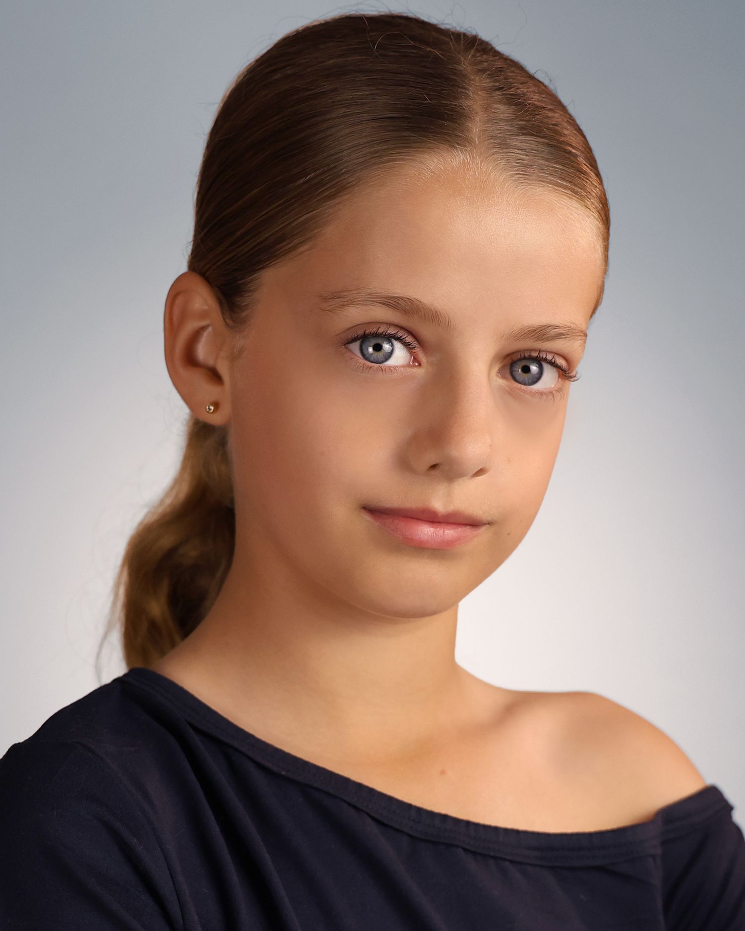 Young girl with blue eyes and braided hair wearing a navy off-shoulder top poses for a professional headshot.