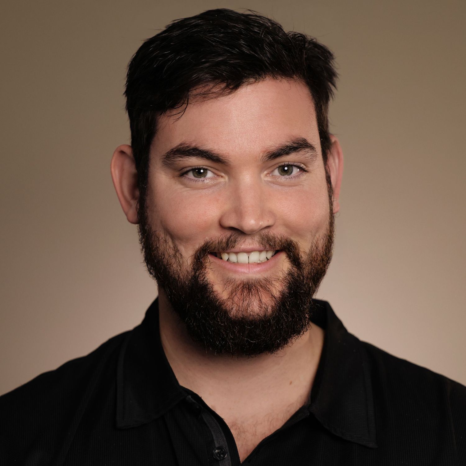 Smiling man with dark beard and blue eyes wearing a black shirt against a neutral brown background.