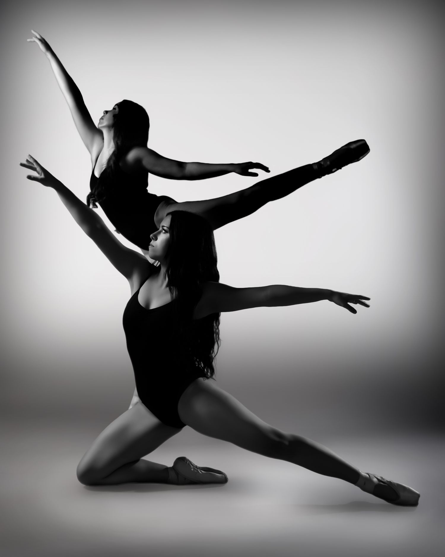 Two female dancers pose gracefully in black leotards, captured in a dramatic black and white studio photograph.
