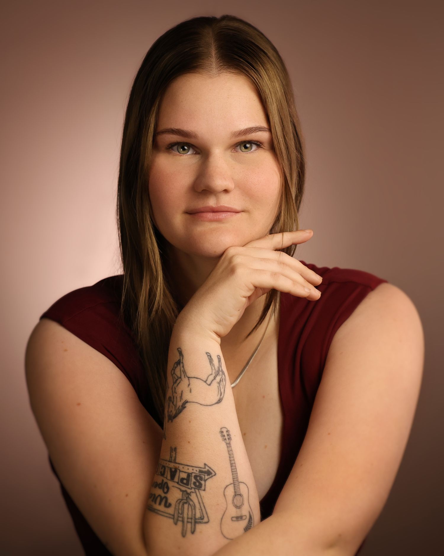 Young woman with long hair and tattoos on her forearm poses confidently in a maroon top against a warm background.