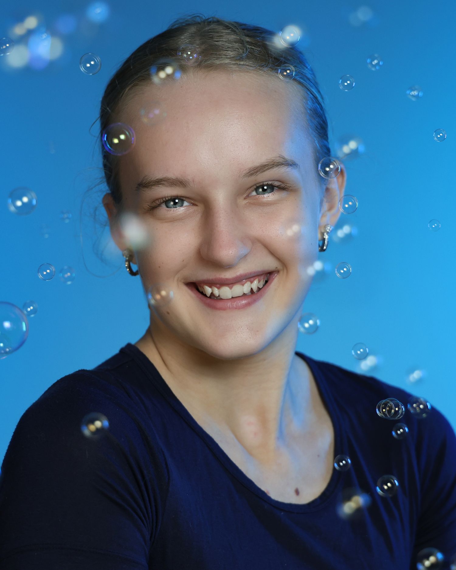 Smiling teenage girl in navy blue top poses for portrait photo with floating soap bubbles on blue background.