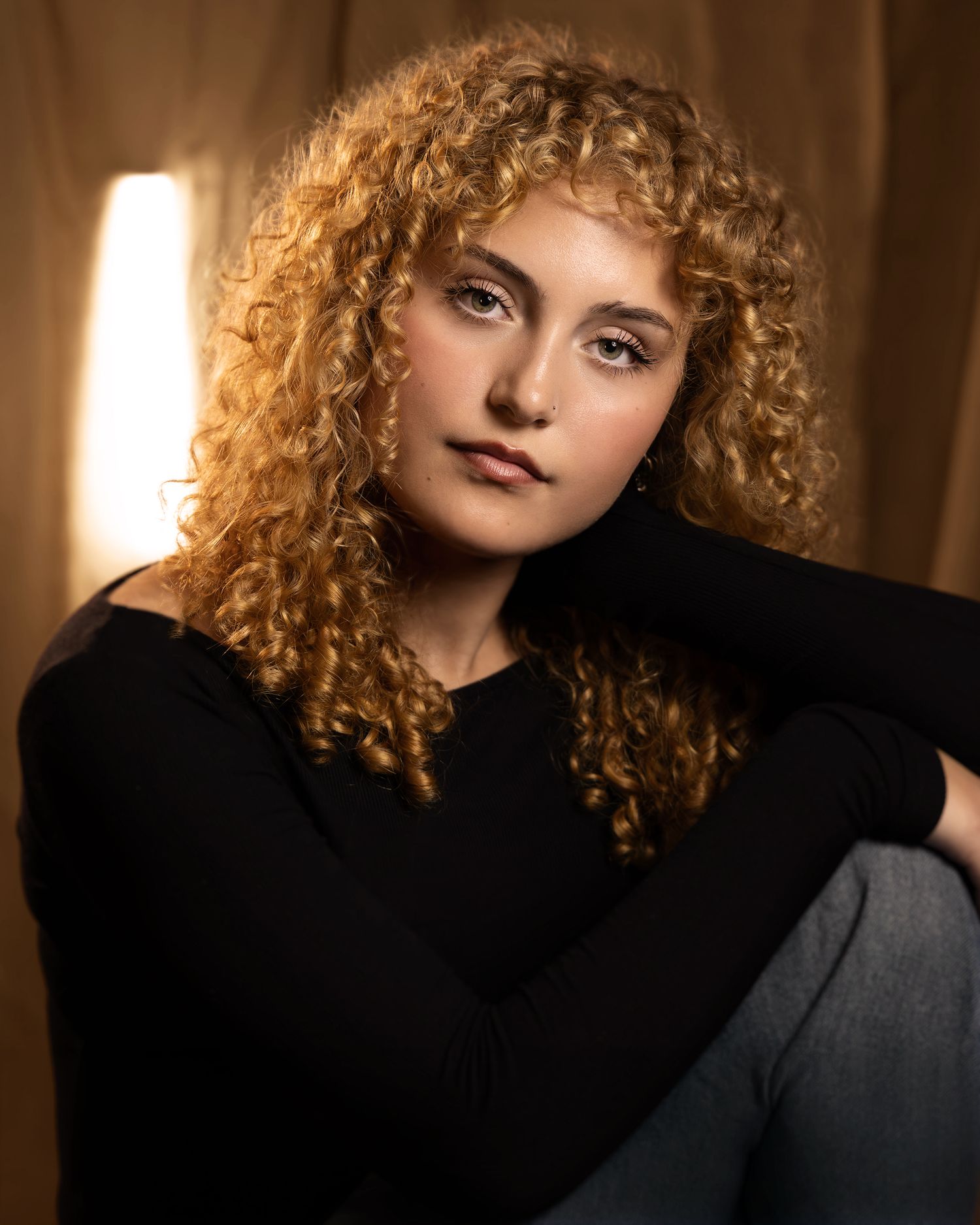 Young woman with curly auburn hair wearing a black top, posed thoughtfully against a warm golden background.