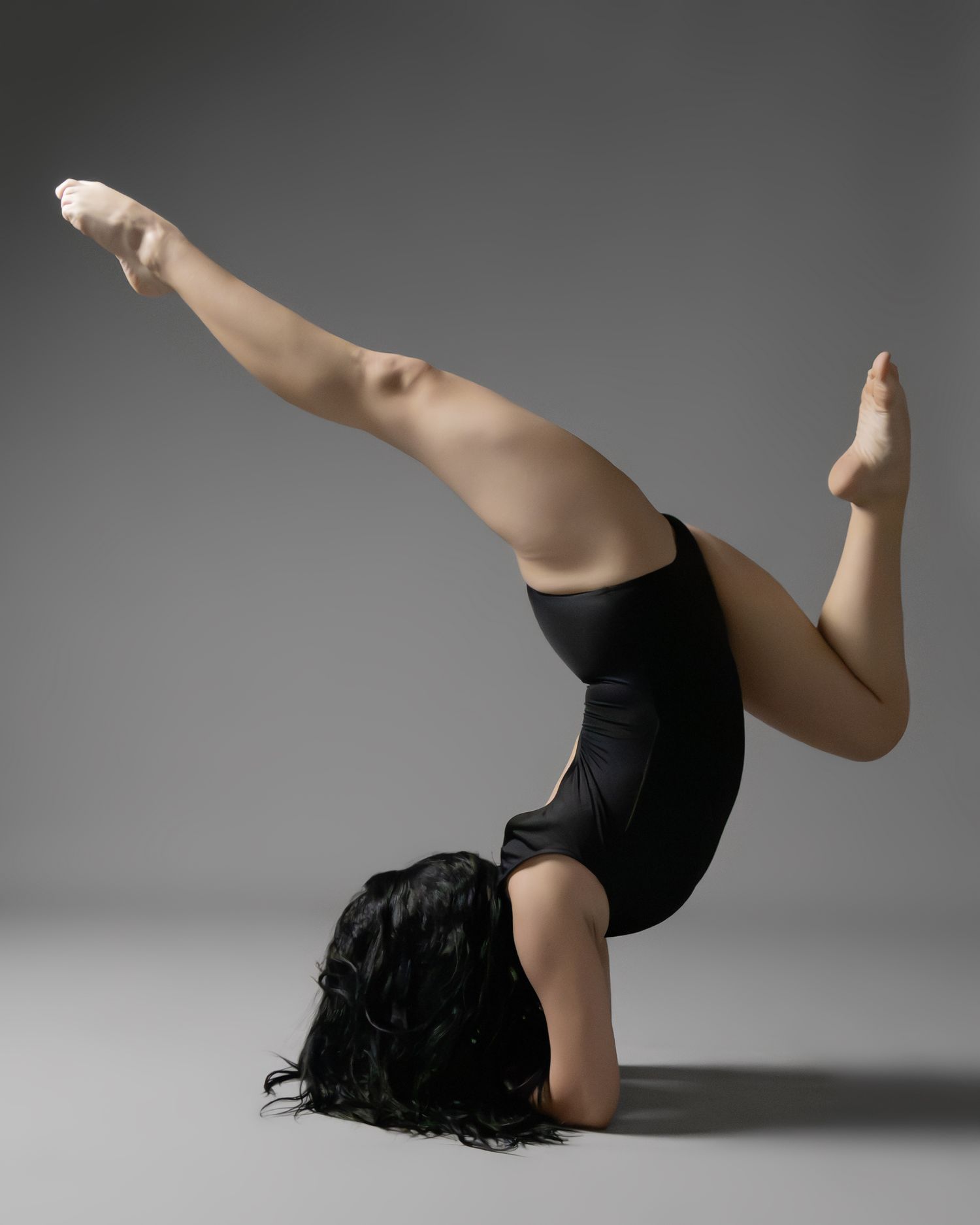 Flexible dancer performing a headstand split in a black leotard against a gray studio background.