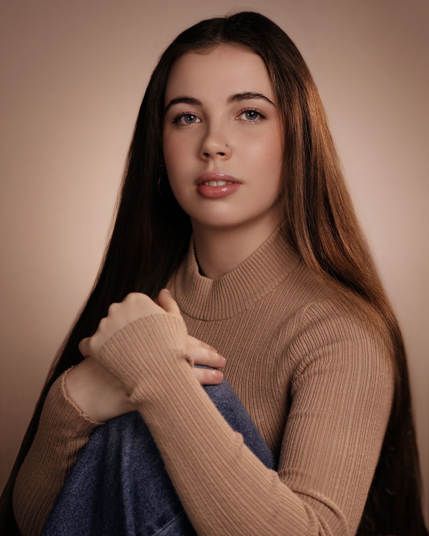 Young woman with long brown hair in a beige ribbed turtleneck sweater holding a blue scarf against neutral background.