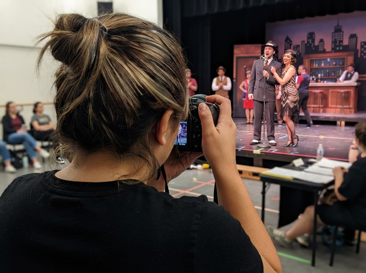 Photographer capturing actors performing on stage during a theater rehearsal in a school auditorium.