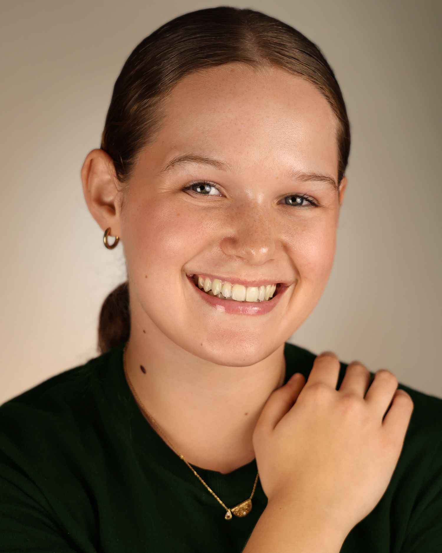 Young woman with freckles smiling warmly, wearing a green top and gold necklace, hair pulled back.