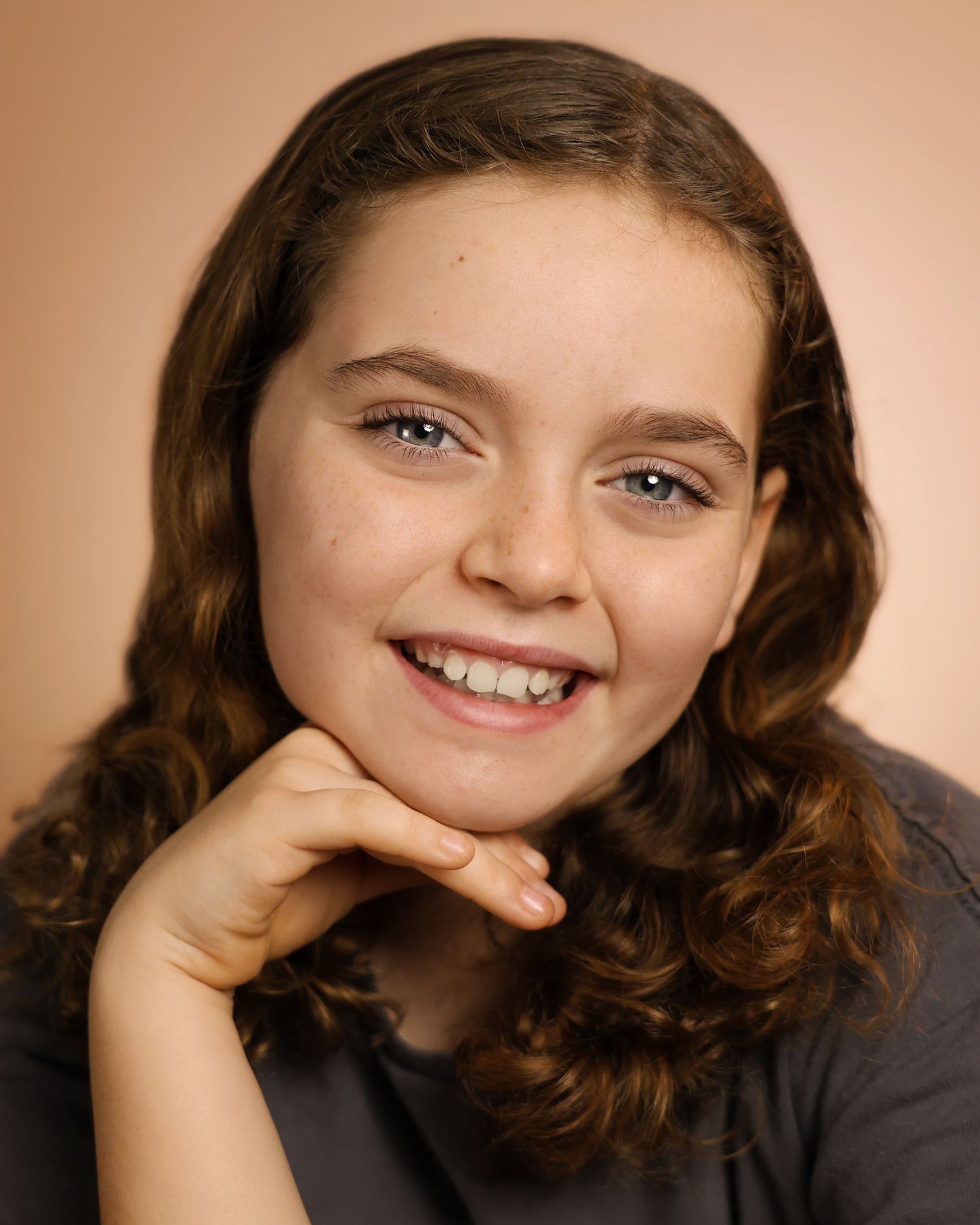 Smiling young girl with curly brown hair resting chin on hand against a warm beige studio background.