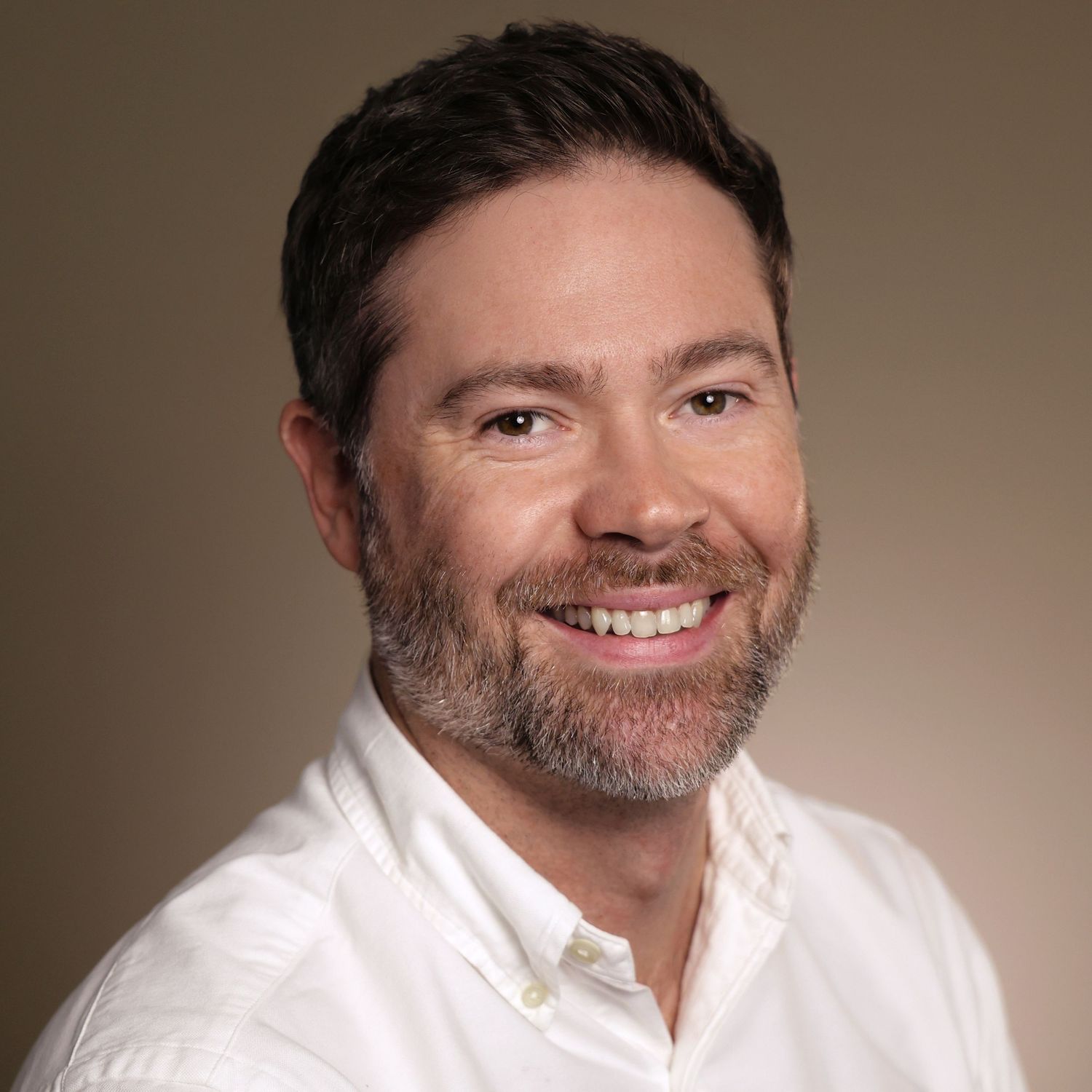 Professional headshot of a smiling man with dark hair and beard wearing a white shirt against a neutral background.