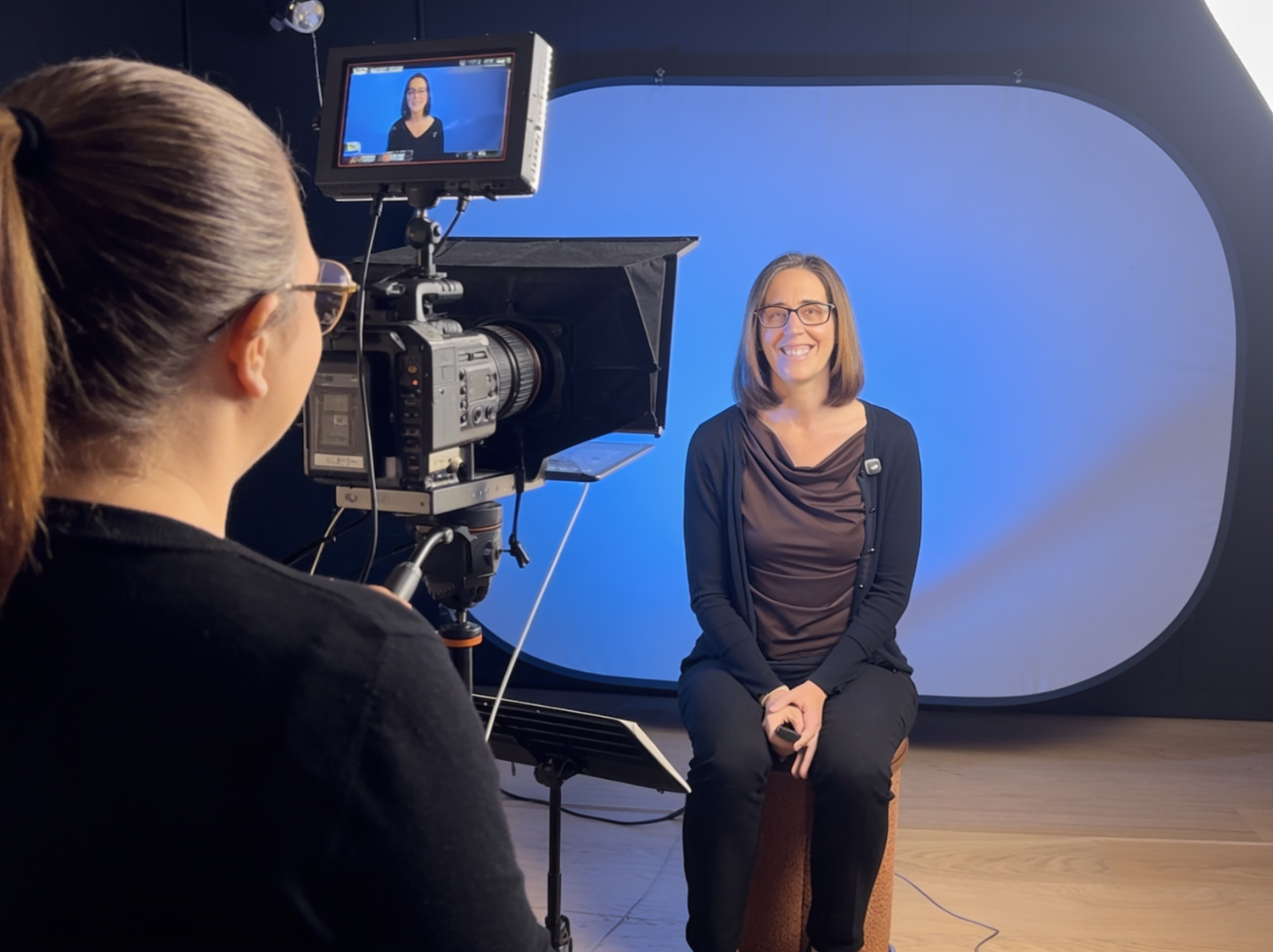Woman being filmed in a professional TV studio setup with a camera operator and blue backdrop.