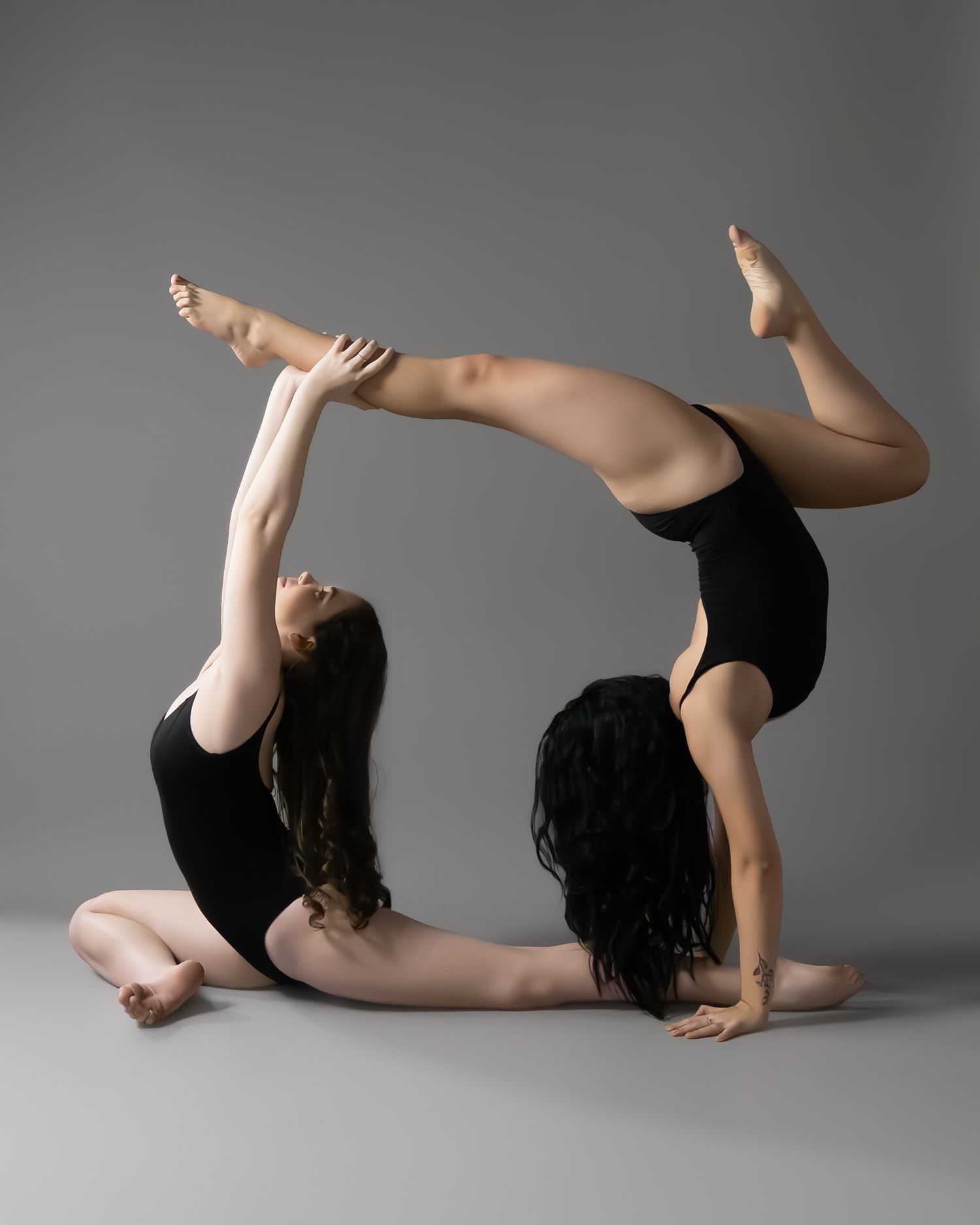 Two flexible dancers in black leotards perform an acrobatic pose together on a gray studio background.