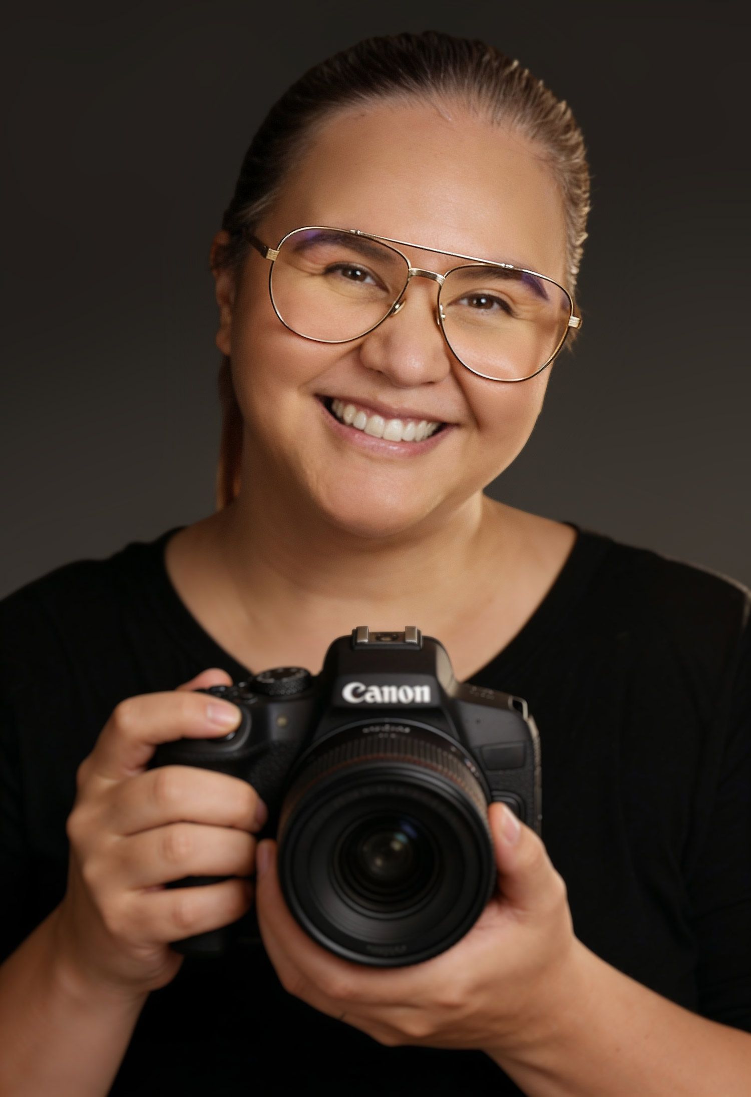 Smiling woman with glasses holding a Canon DSLR camera, wearing a black top against a dark studio background.
