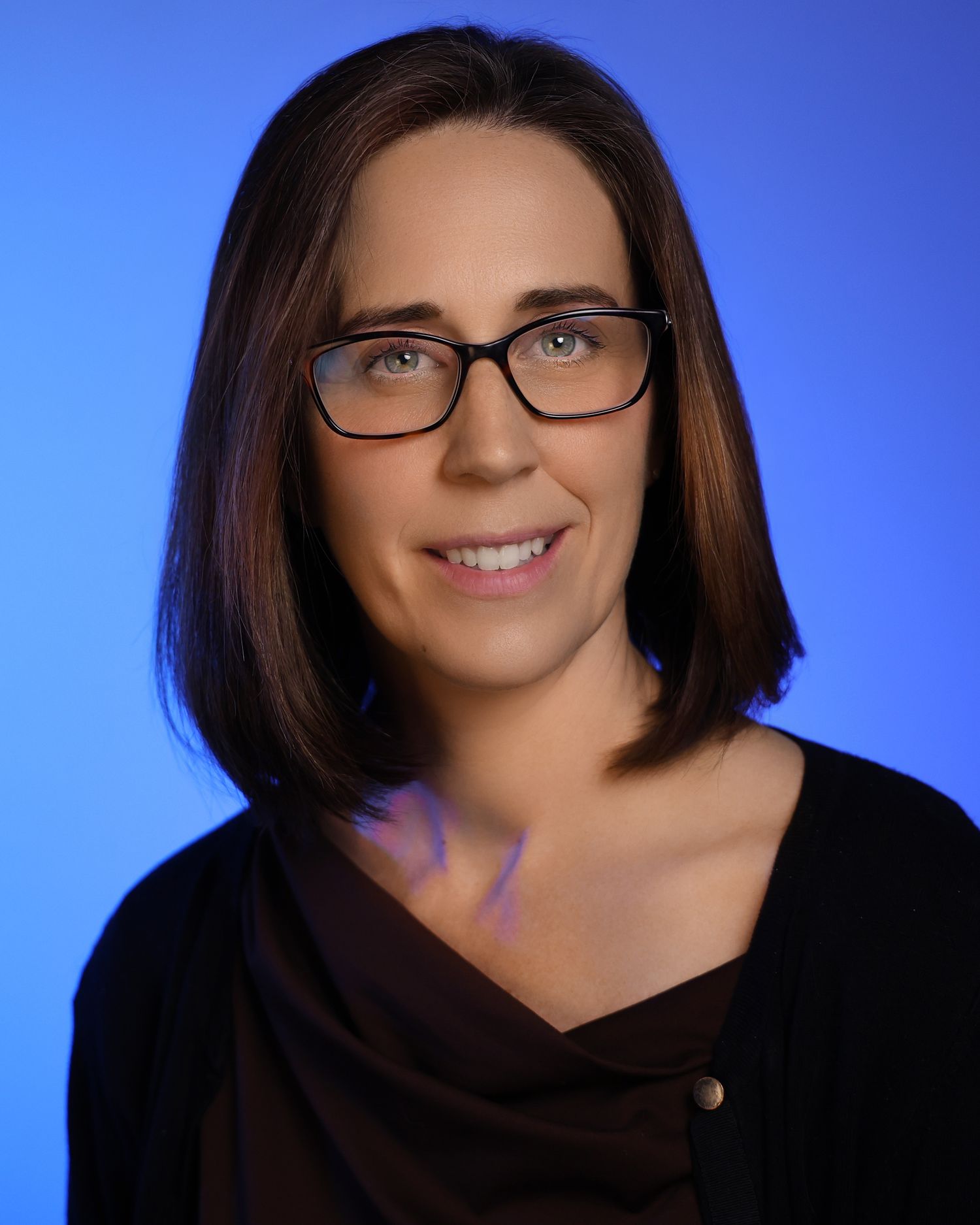 Professional headshot of a woman with dark hair and glasses smiling against a blue background.