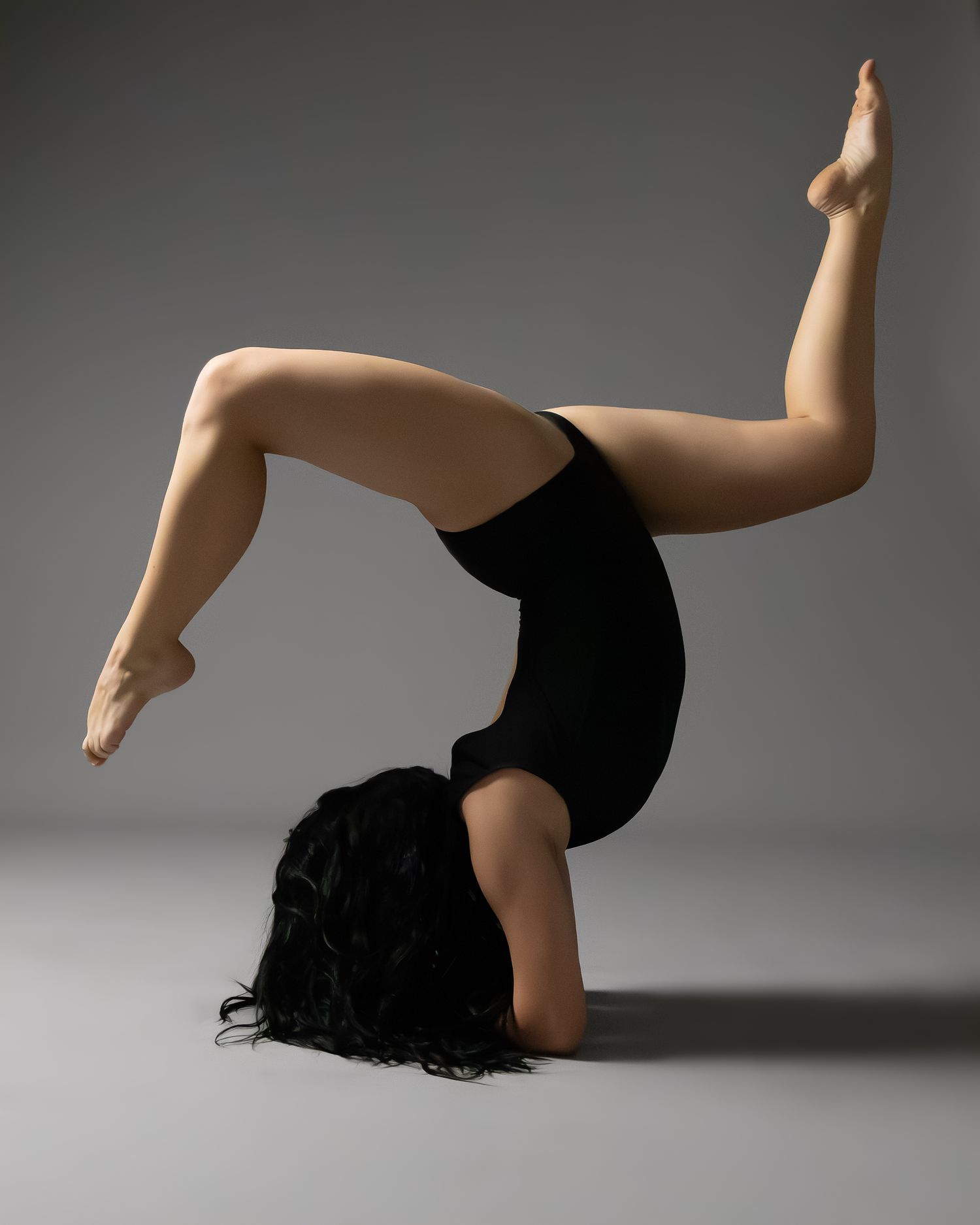 Flexible gymnast in black leotard performing a backbend with legs raised on gray studio background.