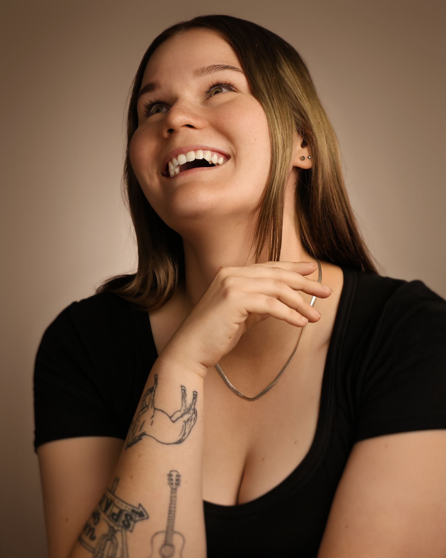 Smiling young woman with tattoos on her arm, wearing a black top, laughing joyfully against a warm brown background.