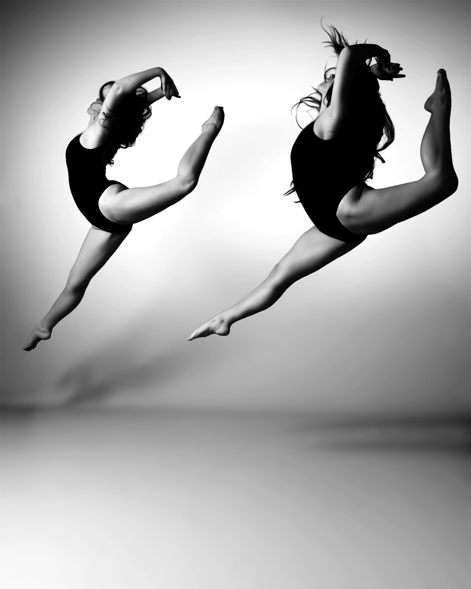 Two female dancers leap gracefully in black leotards, captured in a stunning black and white mid-air dance photography shot.