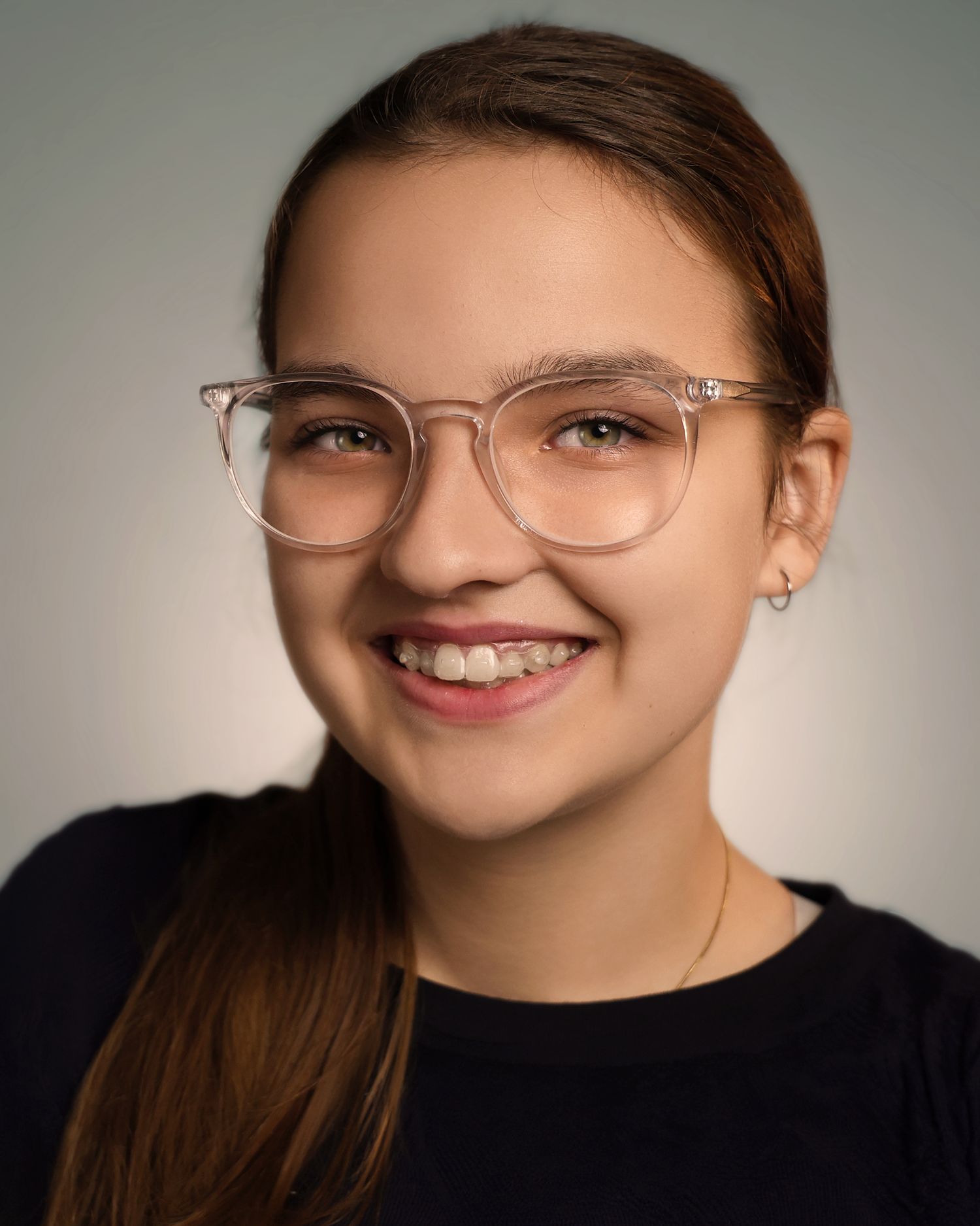 Smiling teenage girl with clear-framed glasses, braces, and long brown hair wearing a black top against a gray background.
