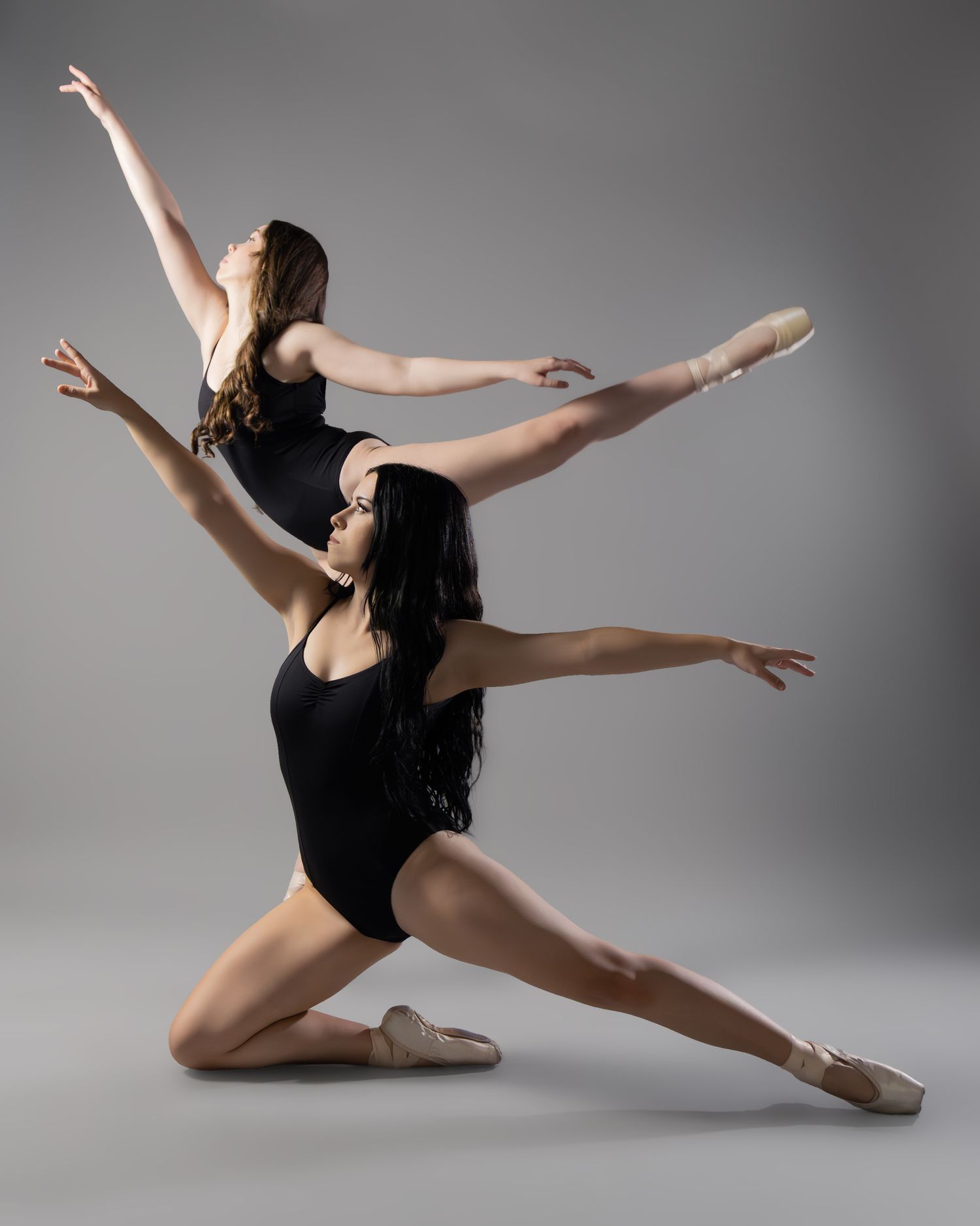 Two ballet dancers in black leotards perform an elegant contemporary pose against a gray studio background.