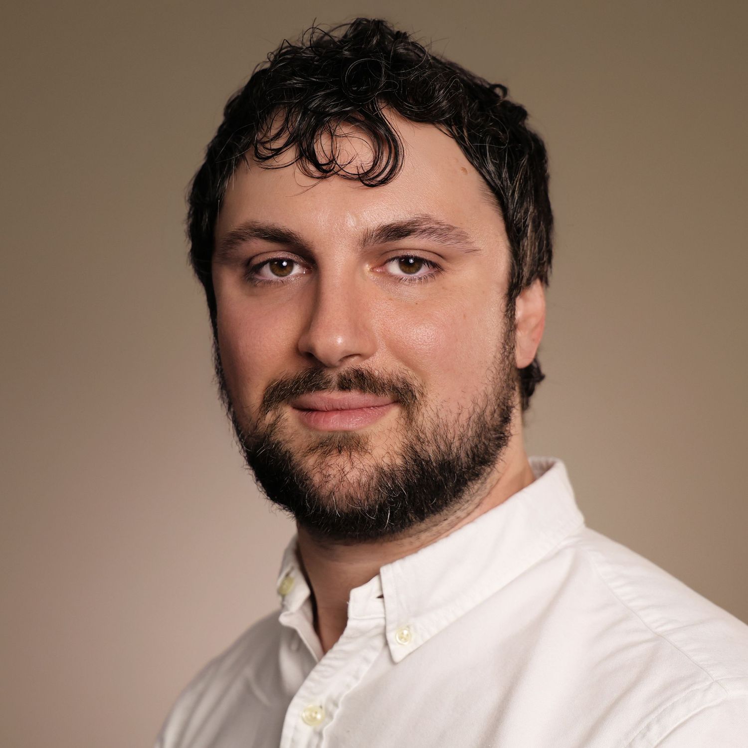 Professional headshot of a bearded man with dark curly hair wearing a white shirt against a neutral background.
