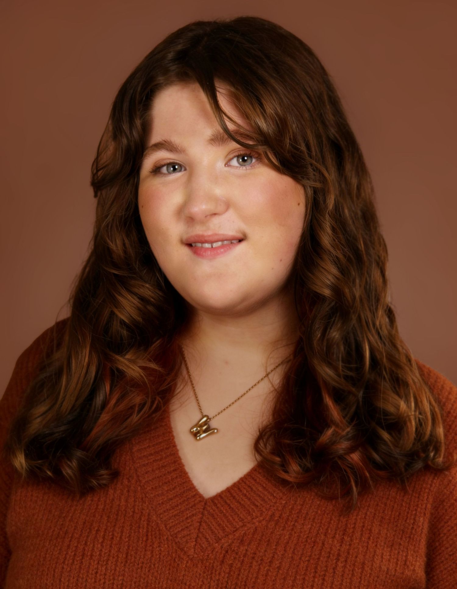 Young woman with curly auburn hair wearing an orange sweater and gold necklace, smiling in a studio portrait.