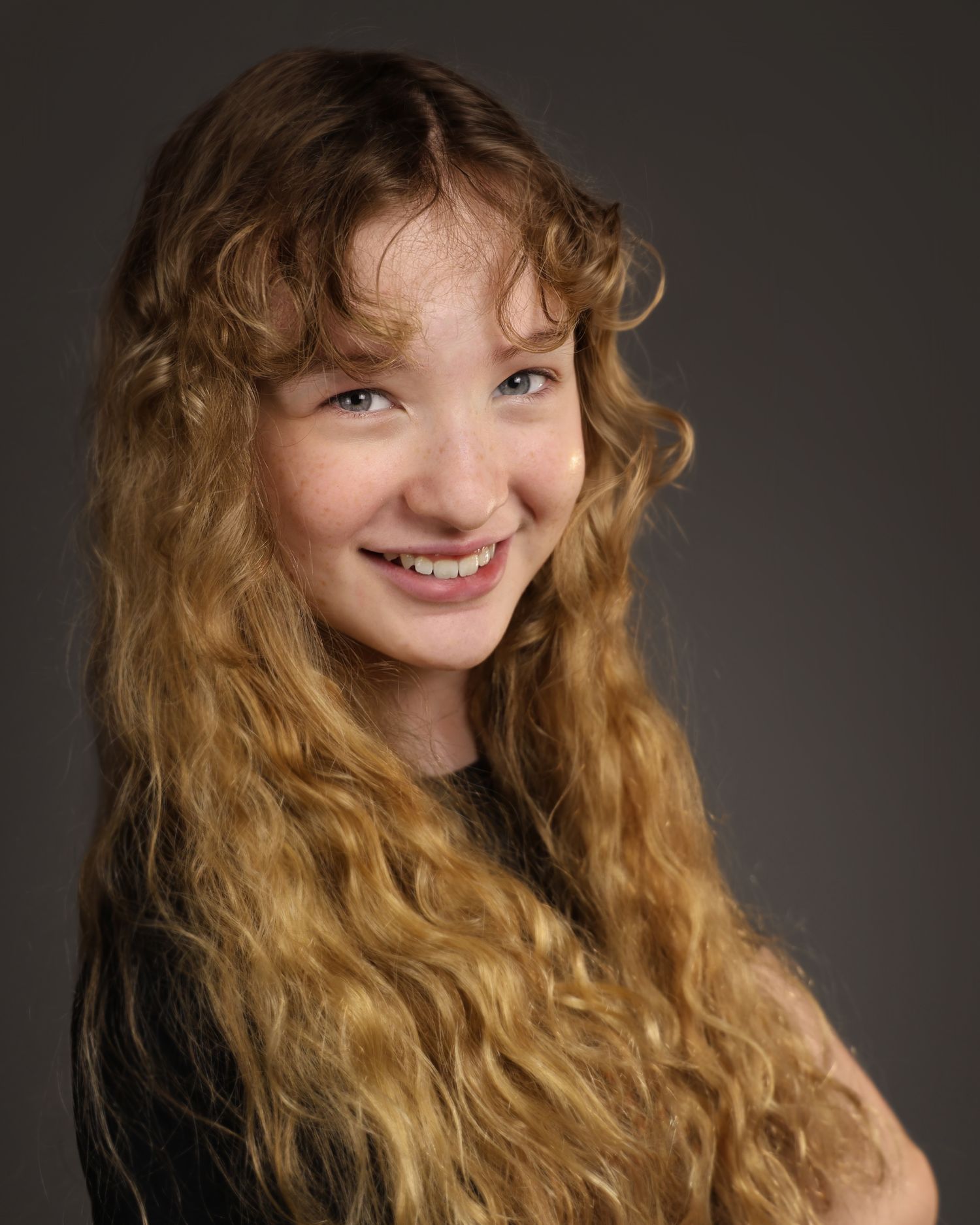 Young girl with long curly auburn hair smiling confidently against a gray studio background.