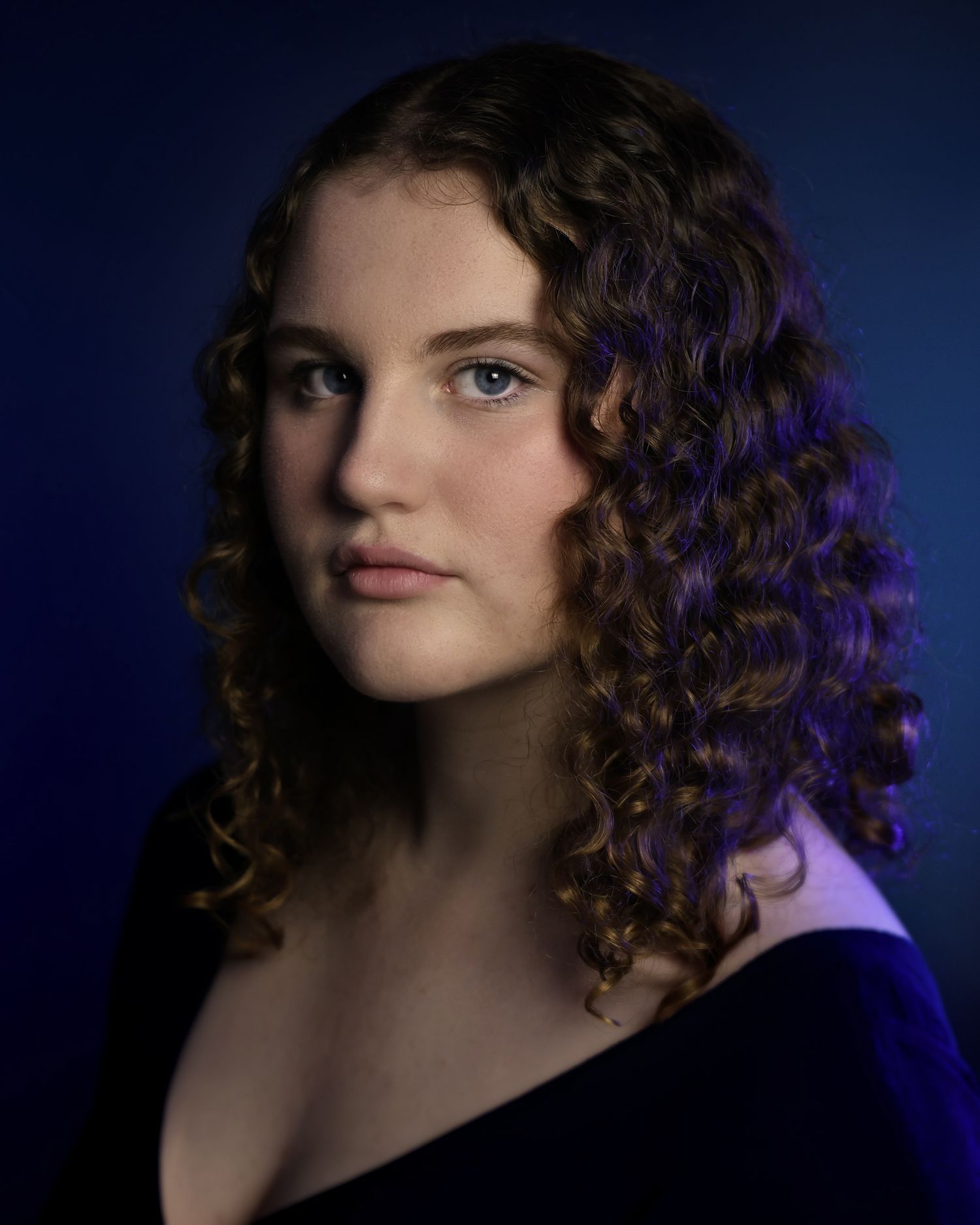 Young woman with curly brown hair and blue eyes poses against a dark blue background in a professional portrait.