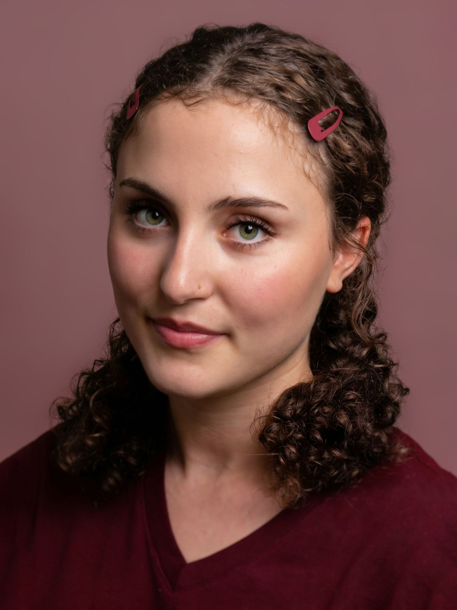 Young woman with curly brown hair and red hair clips wearing burgundy scrubs poses for a professional headshot.