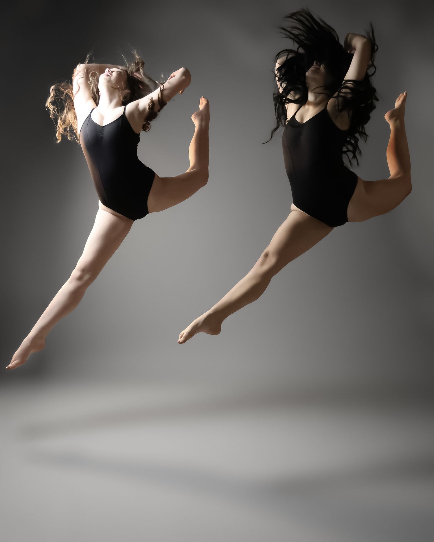 Two female dancers in black leotards leap gracefully mid-air against a gray studio background.