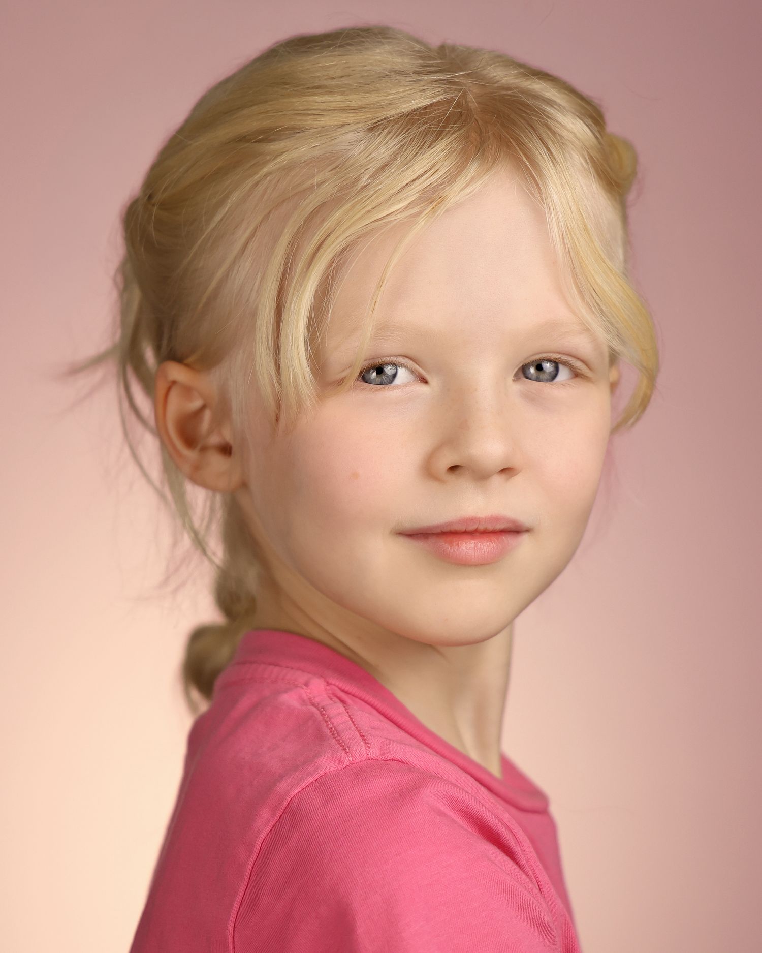 Young blonde girl in a pink shirt poses for a portrait photo against a soft pink background.