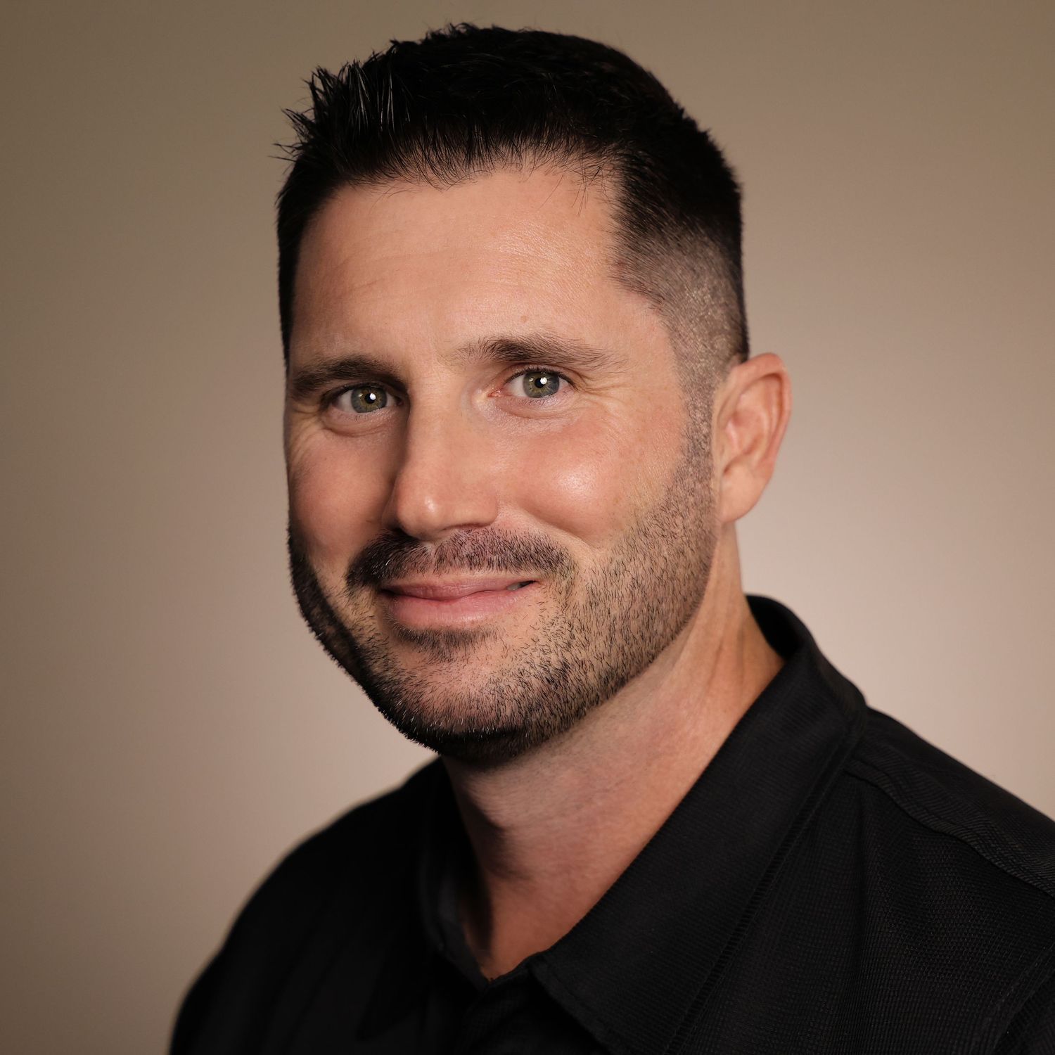 Professional headshot of a smiling man with short dark hair and beard wearing a black shirt against a neutral background.