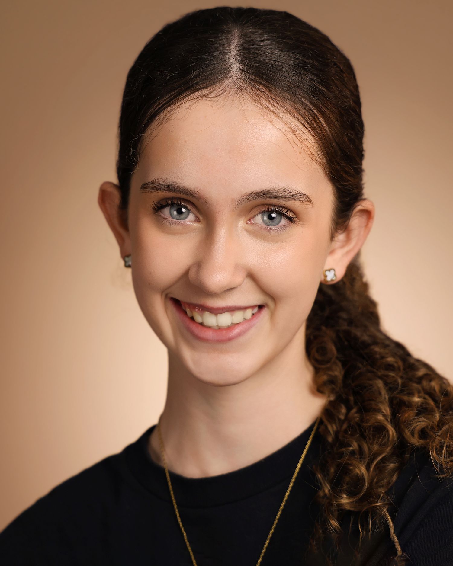 Young woman with curly dark hair, blue eyes, and a warm smile wearing a black top and gold necklace.