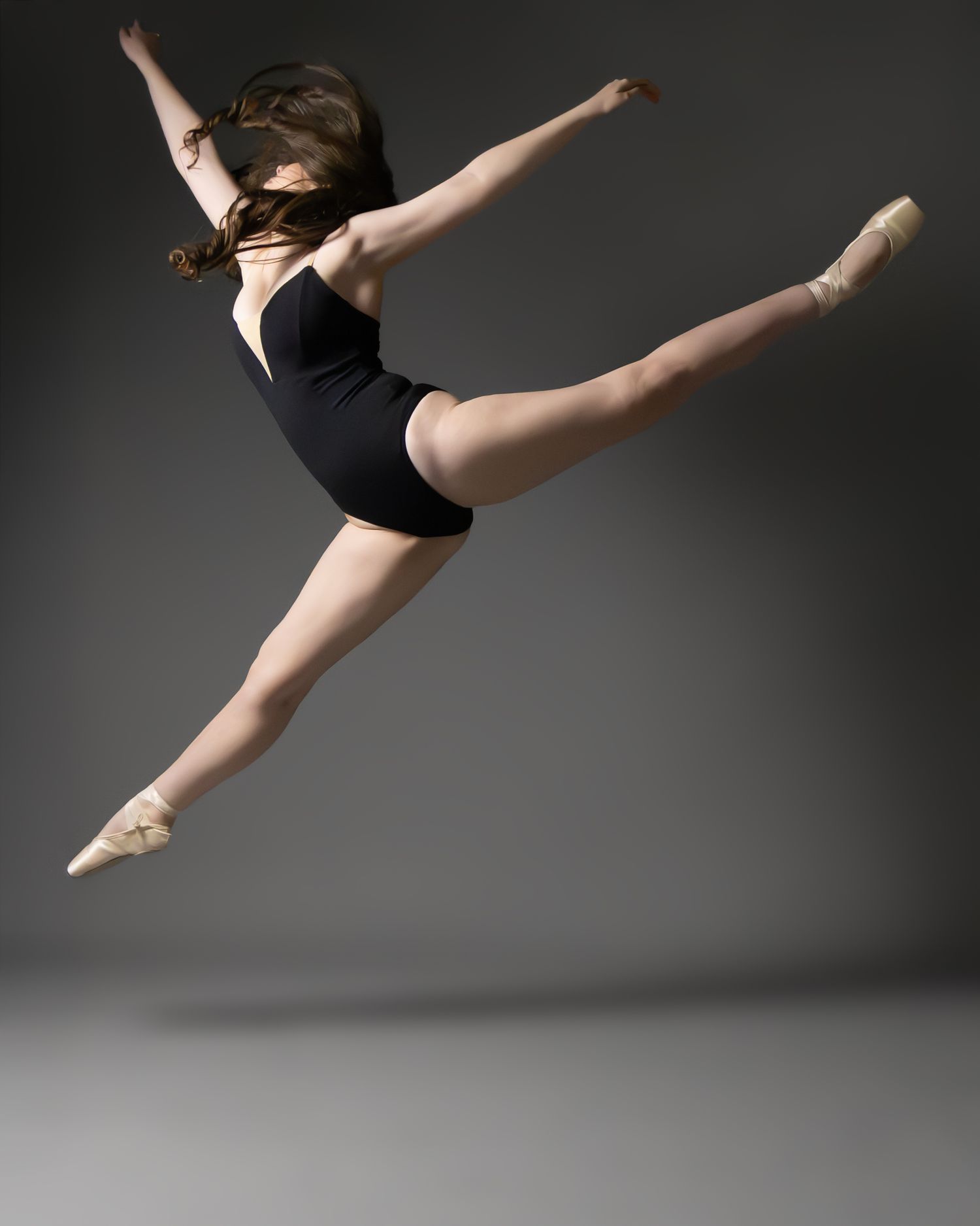 A ballet dancer leaps gracefully in a black leotard and pointe shoes against a gray studio background.