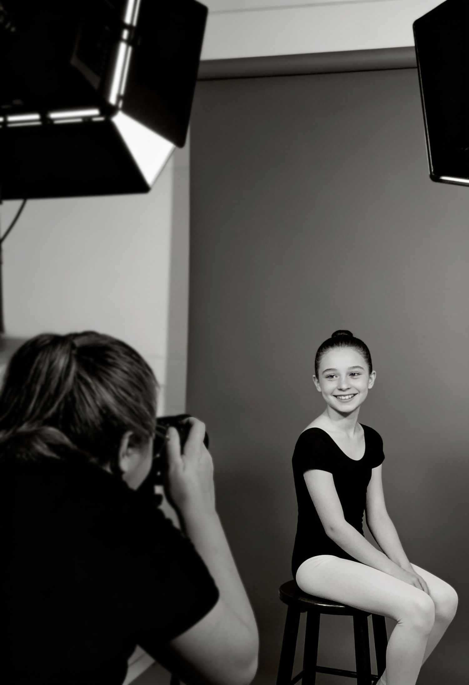 Professional photographer capturing portrait of young girl in black leotard seated on stool in dance studio.