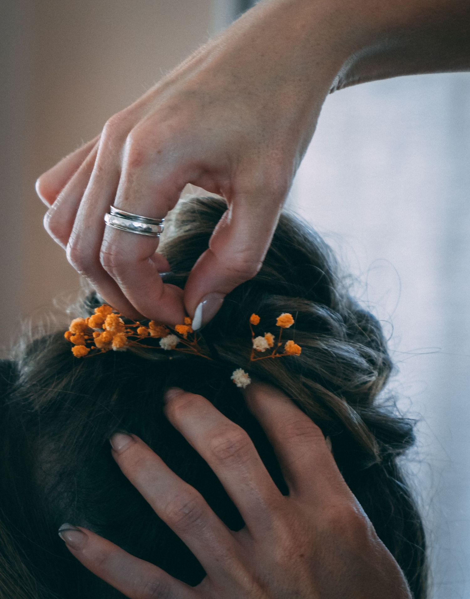 Hands placing small orange flower accents into a bride's dark braided updo hairstyle.