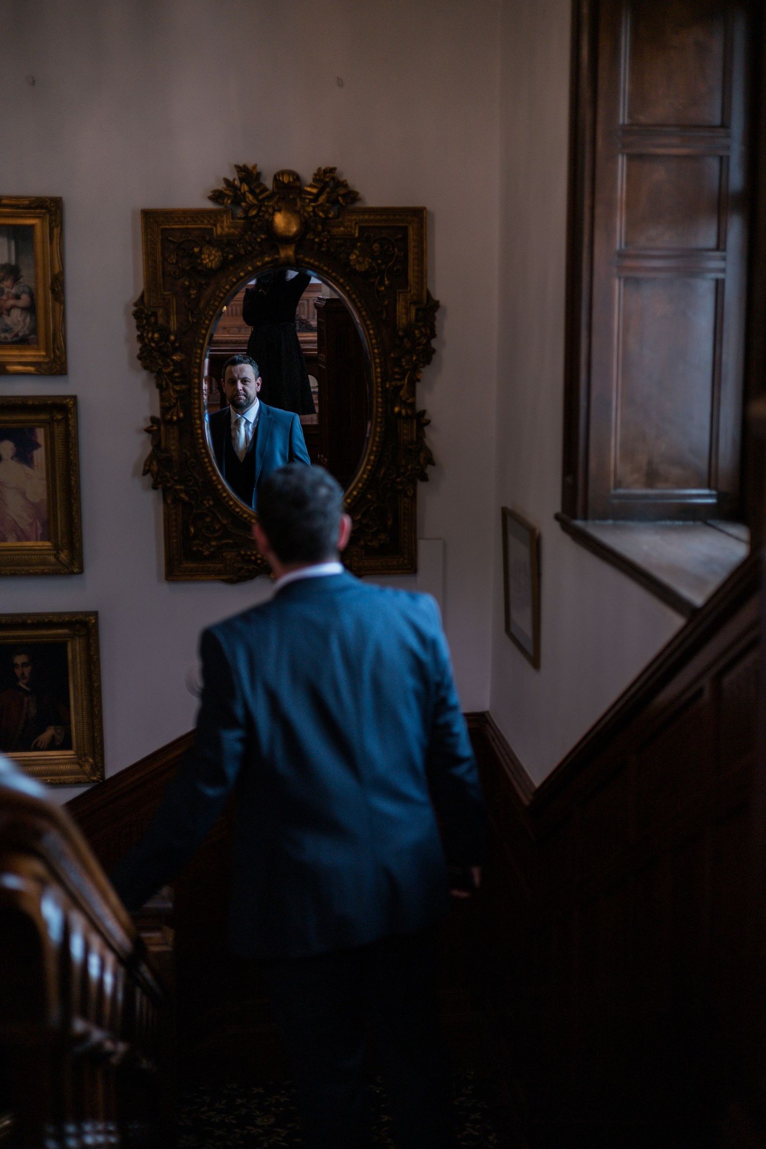 Groom in blue suit ascending staircase reflected in ornate gold mirror at wedding venue.