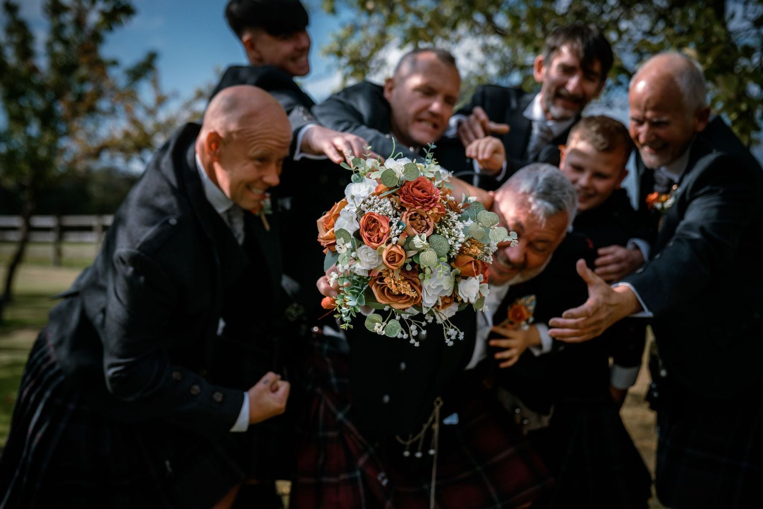Groomsmen in kilts playfully gather around wedding bouquet in fun outdoor photo.