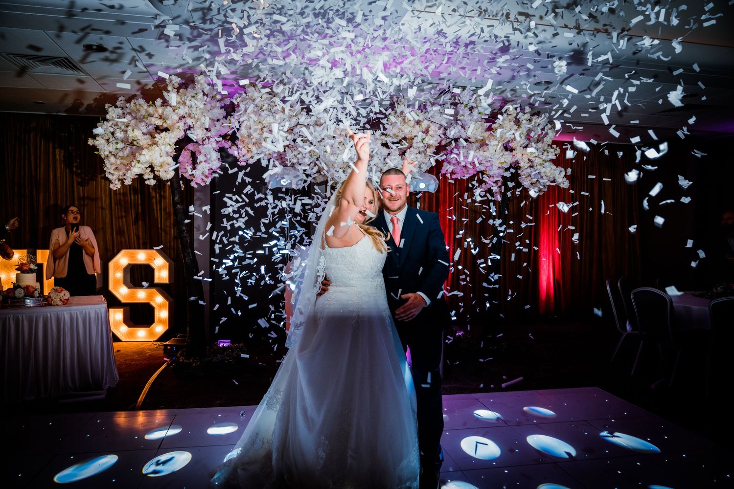 Bride and groom celebrating on dance floor with confetti cannon explosion and illuminated letter decor.