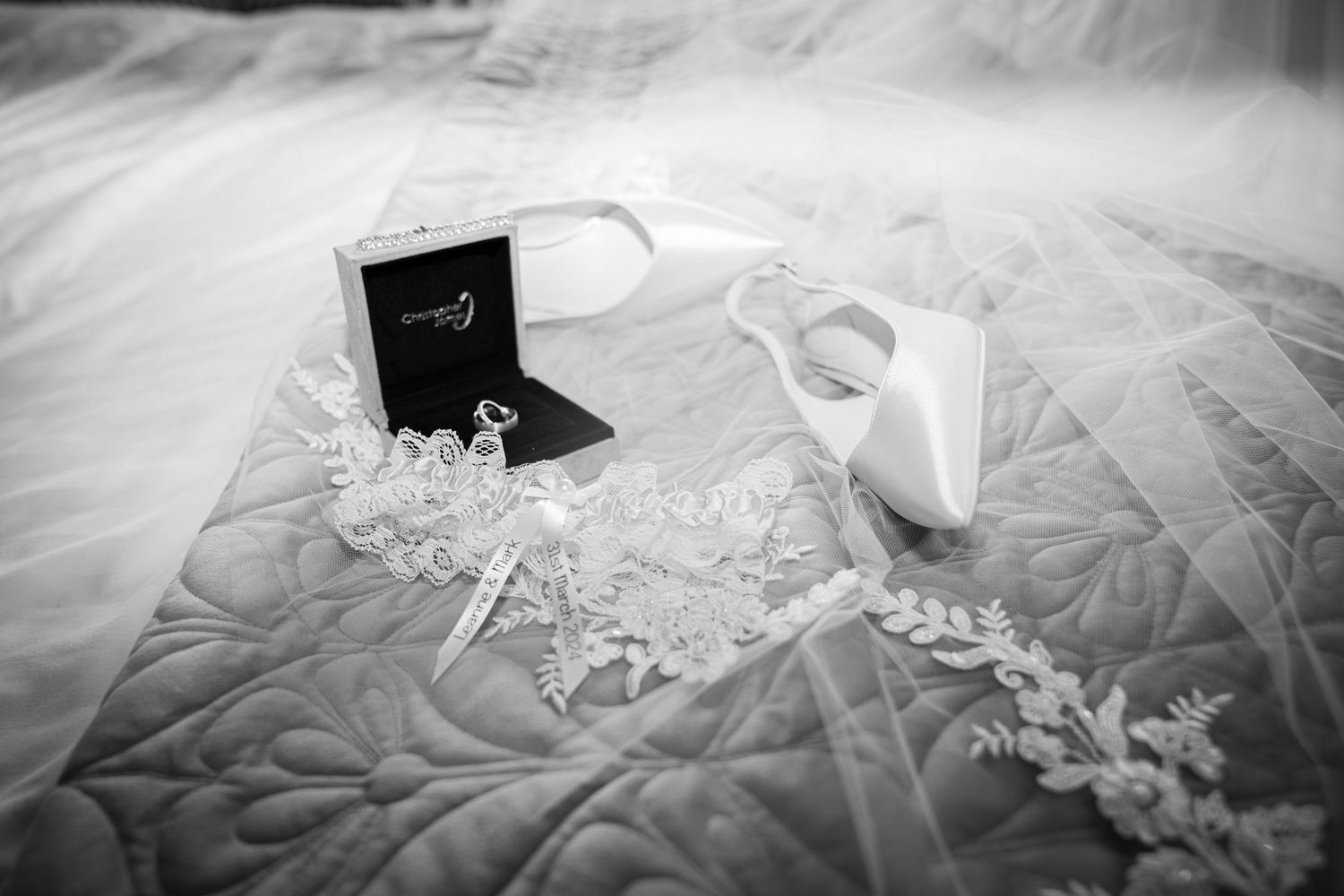 Black and white flat lay of bridal accessories including ring box, heels, and lace veil.